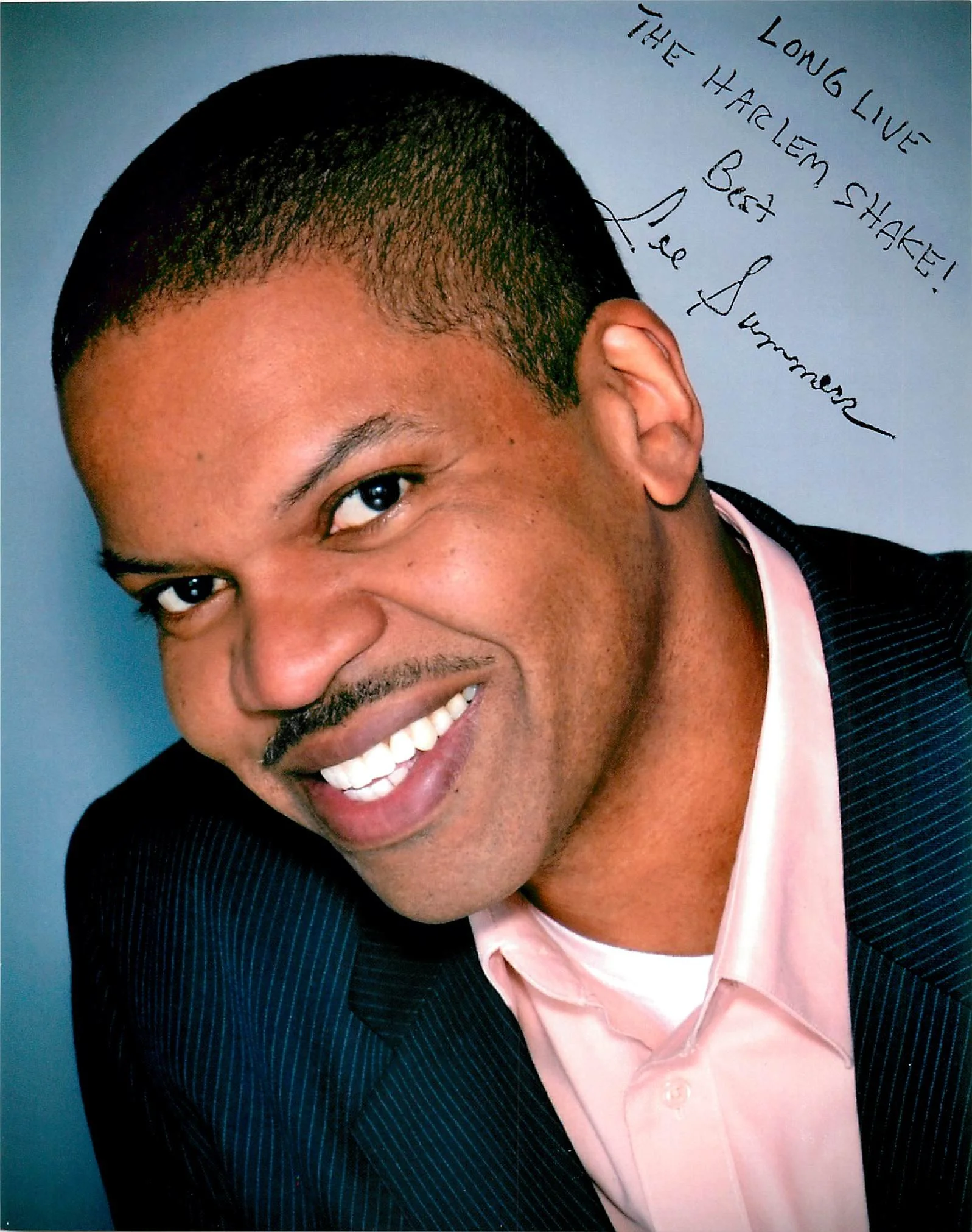 A smiling young man with short black hair, wearing a pink dress shirt and a dark pinstripe blazer, partially visible against a blue background with handwritten text in black ink.