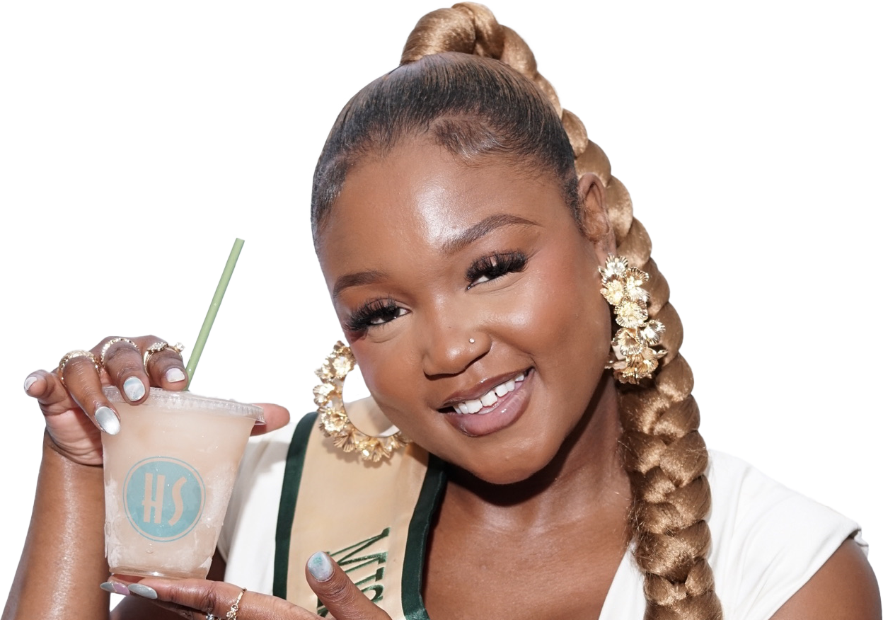 A woman smiling and holding a drink with a straw, wearing large floral earrings, a sash, and with styled hair.