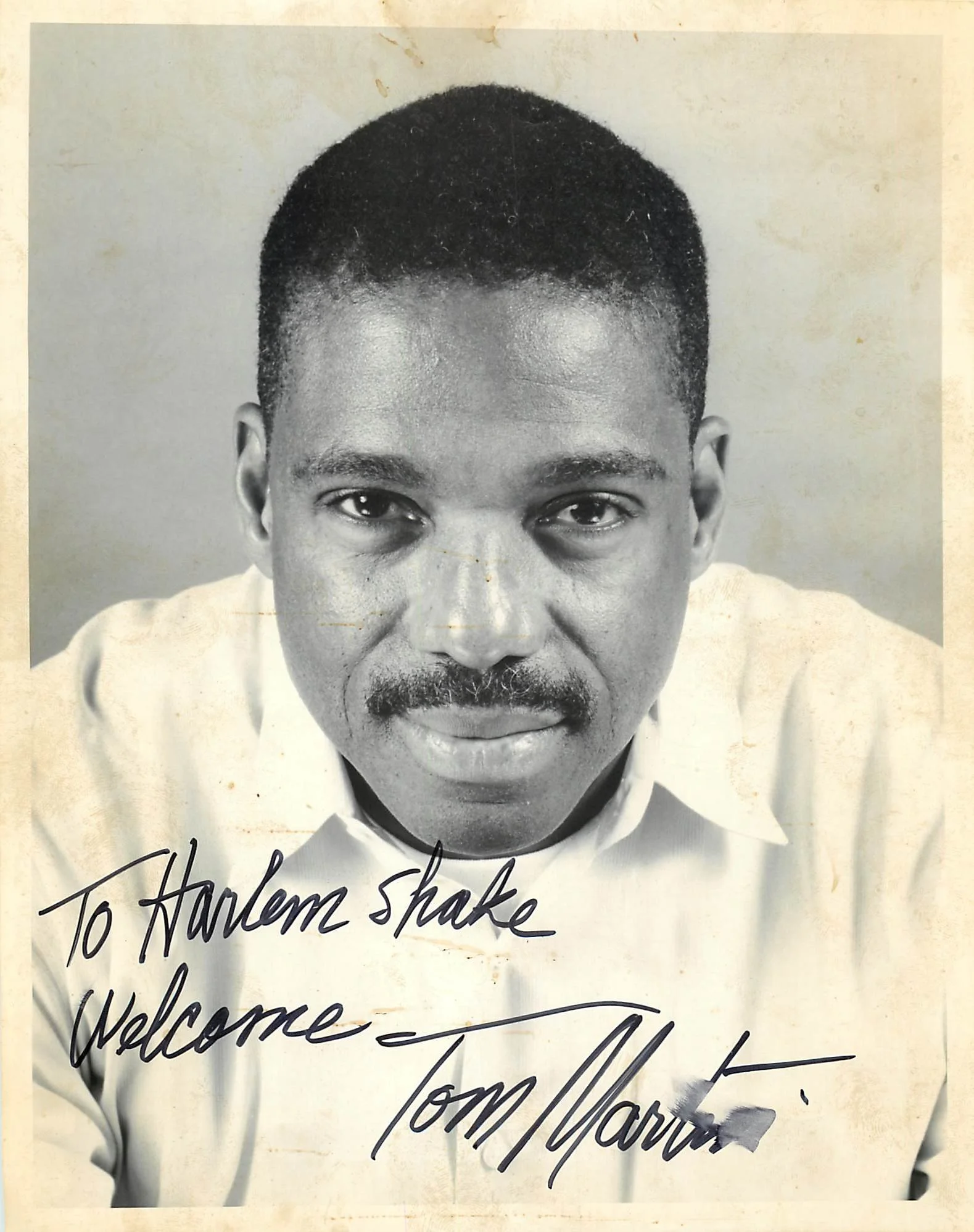 Black and white photograph of a man with short hair and facial hair, wearing a collared shirt. There is handwritten text on the photograph that reads: "To Harlem shake, Welcome - Tom Wallace."