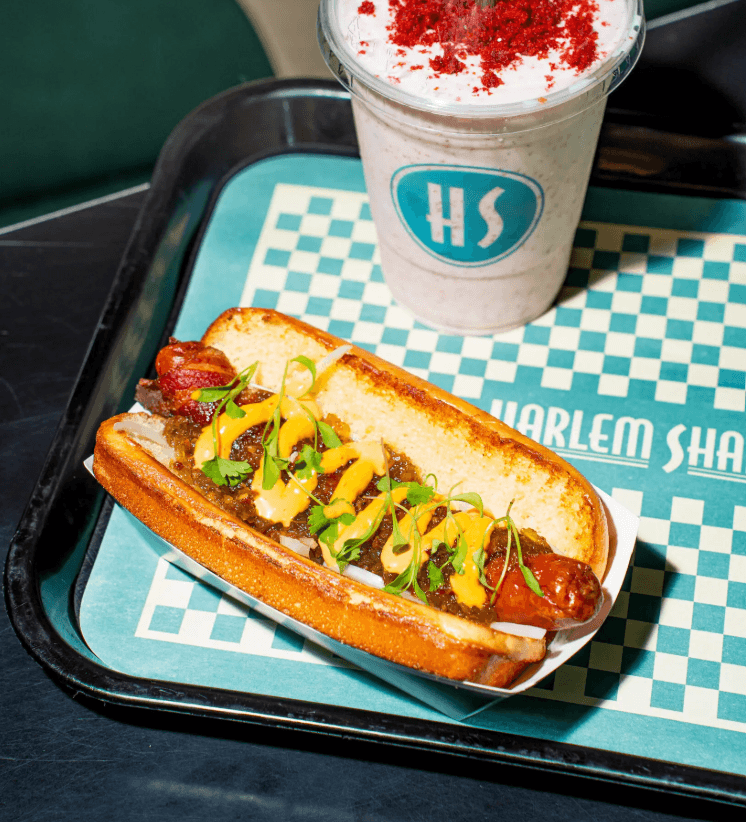 Hot dog with mustard, relish, and microgreens in a toasted bun, served on a tray with a branded drink in a foam cup topped with red seasoning.