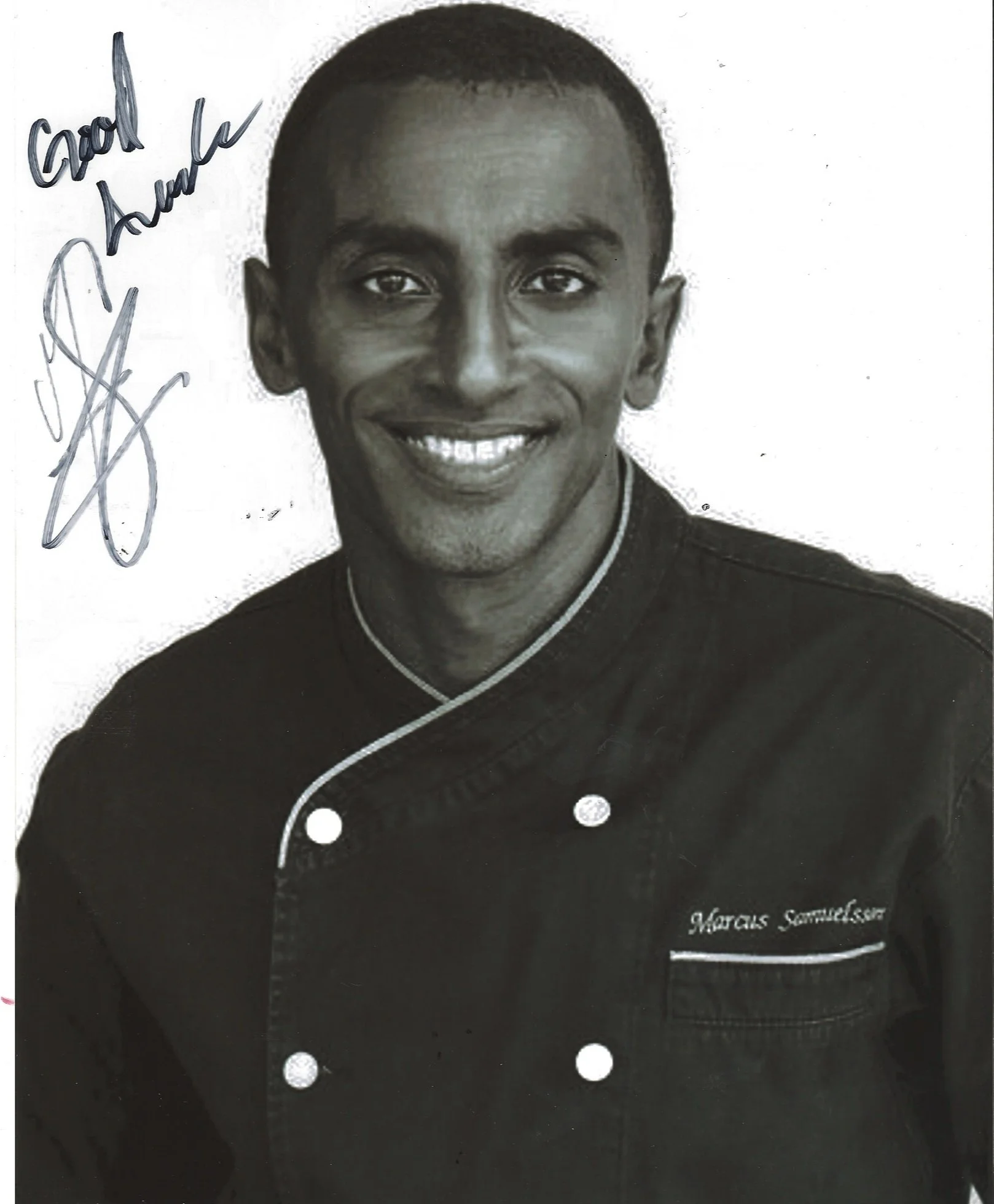 Black and white portrait of Marcus Samuelsson, smiling, wearing a chef's jacket with white piping.