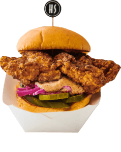 A fried chicken sandwich with pickles, red onions, and a bun, served in a white paper boat with a small flag on top.