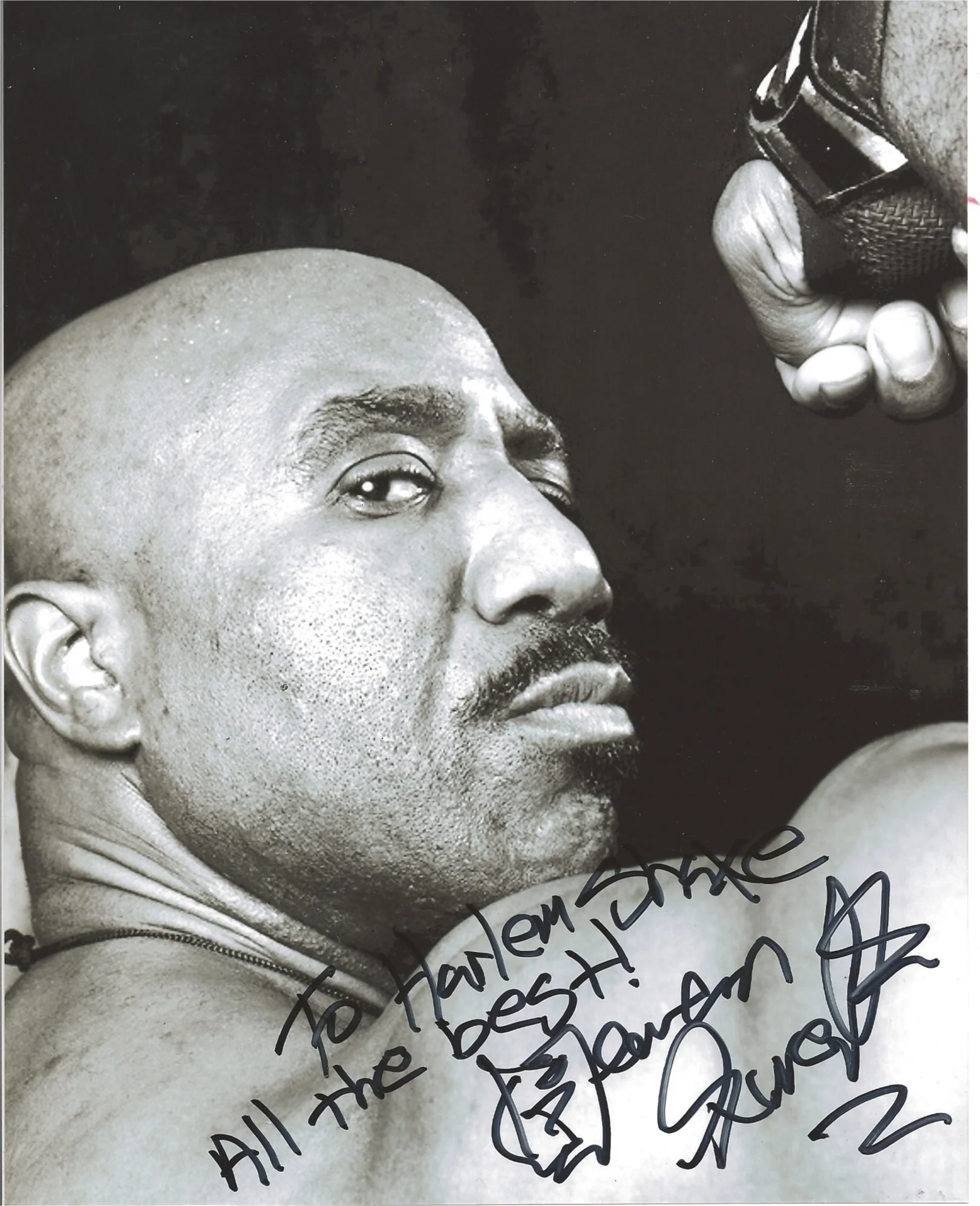 Black and white photograph of a man's face with a shaved head, looking to the side, holding a microphone near his mouth, with handwritten notes or signatures over the bottom part of the image.