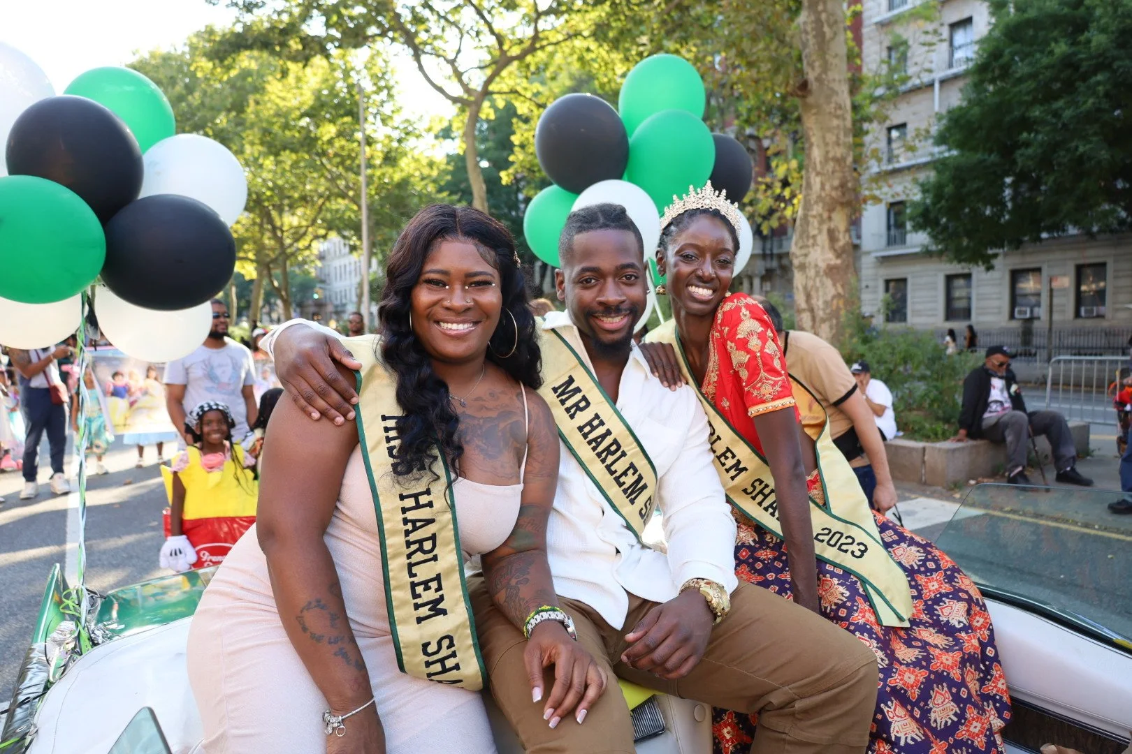 Three individuals sitting on a parade float surrounded by people and balloons, with trees and buildings in the background. They are wearing sashes, and one woman is wearing a crown.