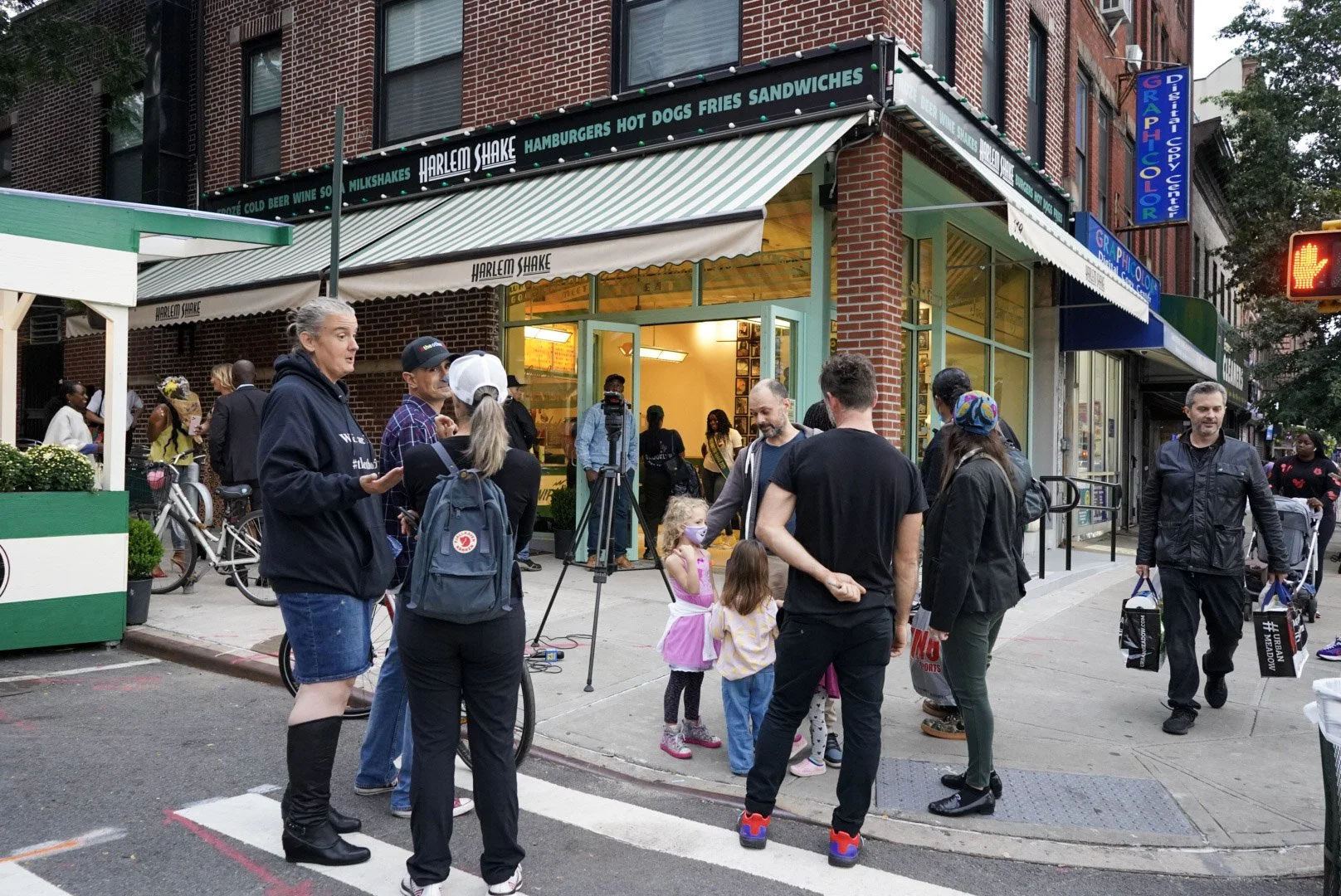 People gathered outside Harlem Shake restaurant on a city street, some talking and children wearing masks, with a camera setup for filming or photography.