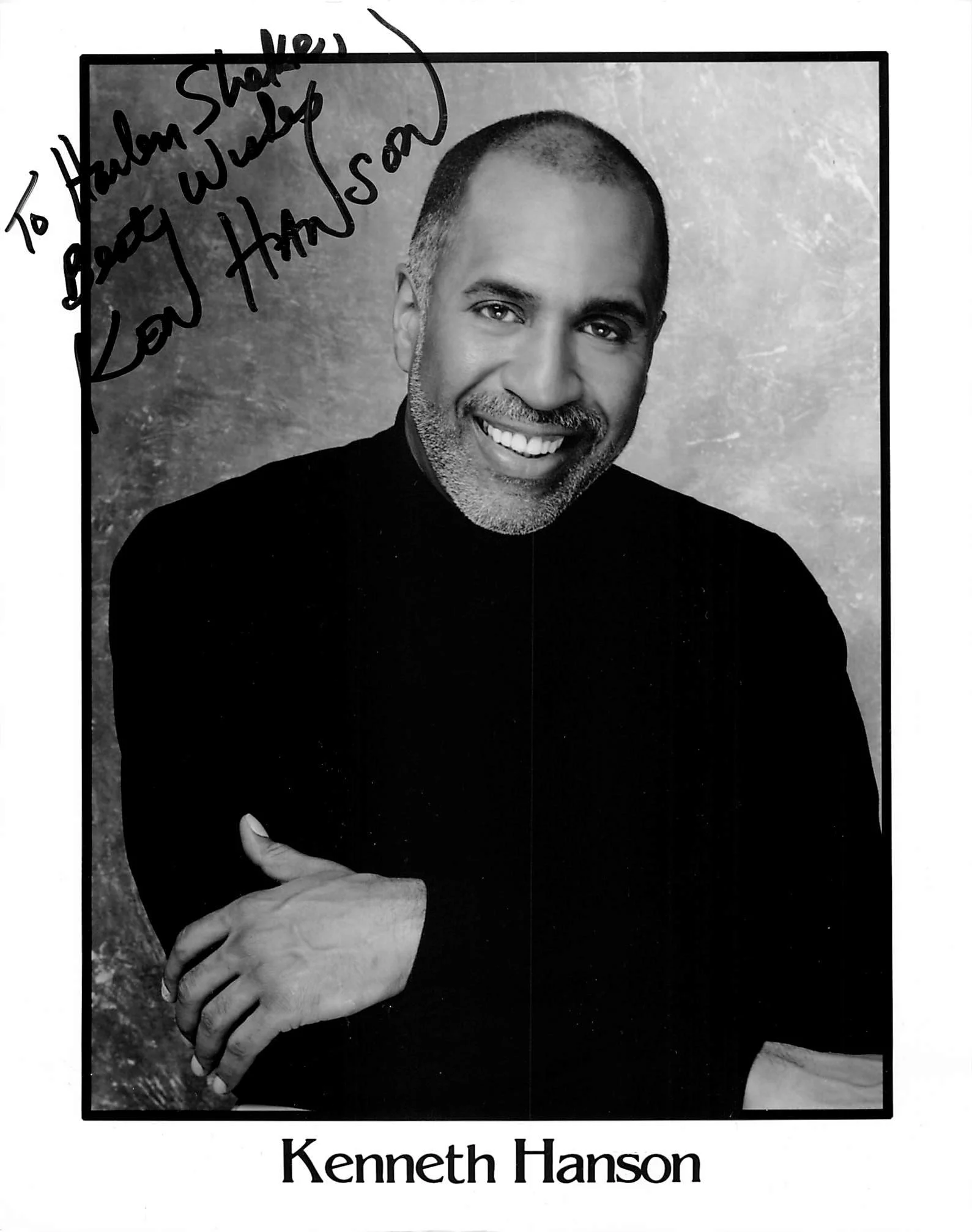 Black and white photograph of a smiling man with short hair and a beard, wearing a black shirt, with his arms crossed. Handwritten text in the top left corner reads, "To Howard Starke, God bless you. Love, Ham on so." The bottom part of the image has