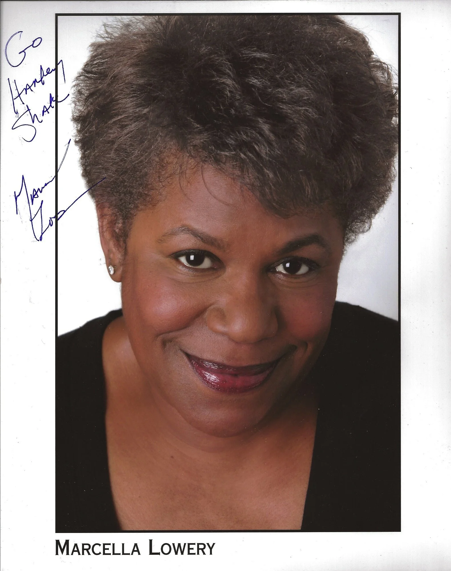 Photograph of a woman with short, curly dark hair and a smile, wearing earrings and a black top. Handwritten note on the left side says, 'Go Harleen, Thank You' in blue ink. Text at the bottom reads, 'Marcella Lowery'.