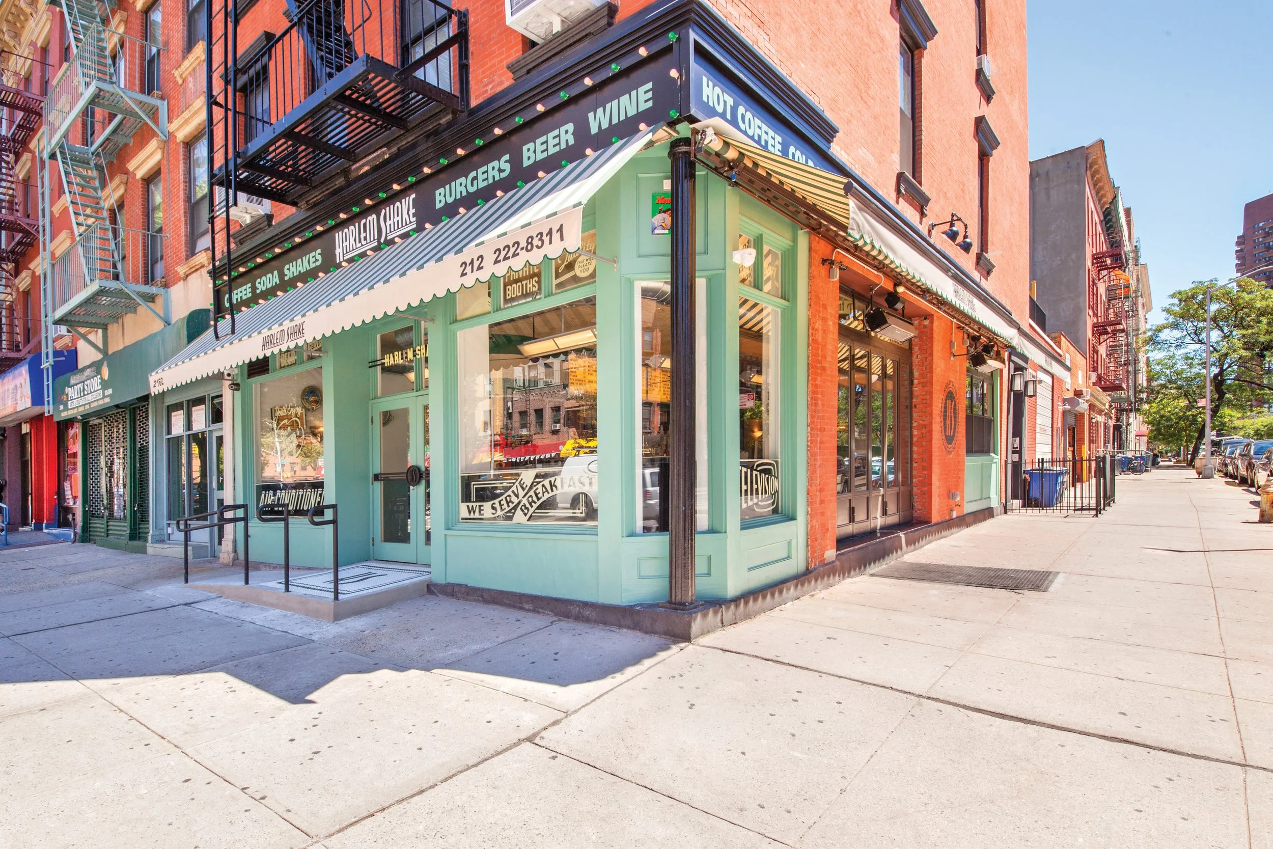 Corner view of a small eatery on a city street, painted mint green with large windows and signs advertising burgers, beer, hot coffee, and cold drinks. The storefront has a striped awning and a phone number displayed. Surrounding buildings are brick with fire escapes, and the street is sunny with some parked cars and trees.