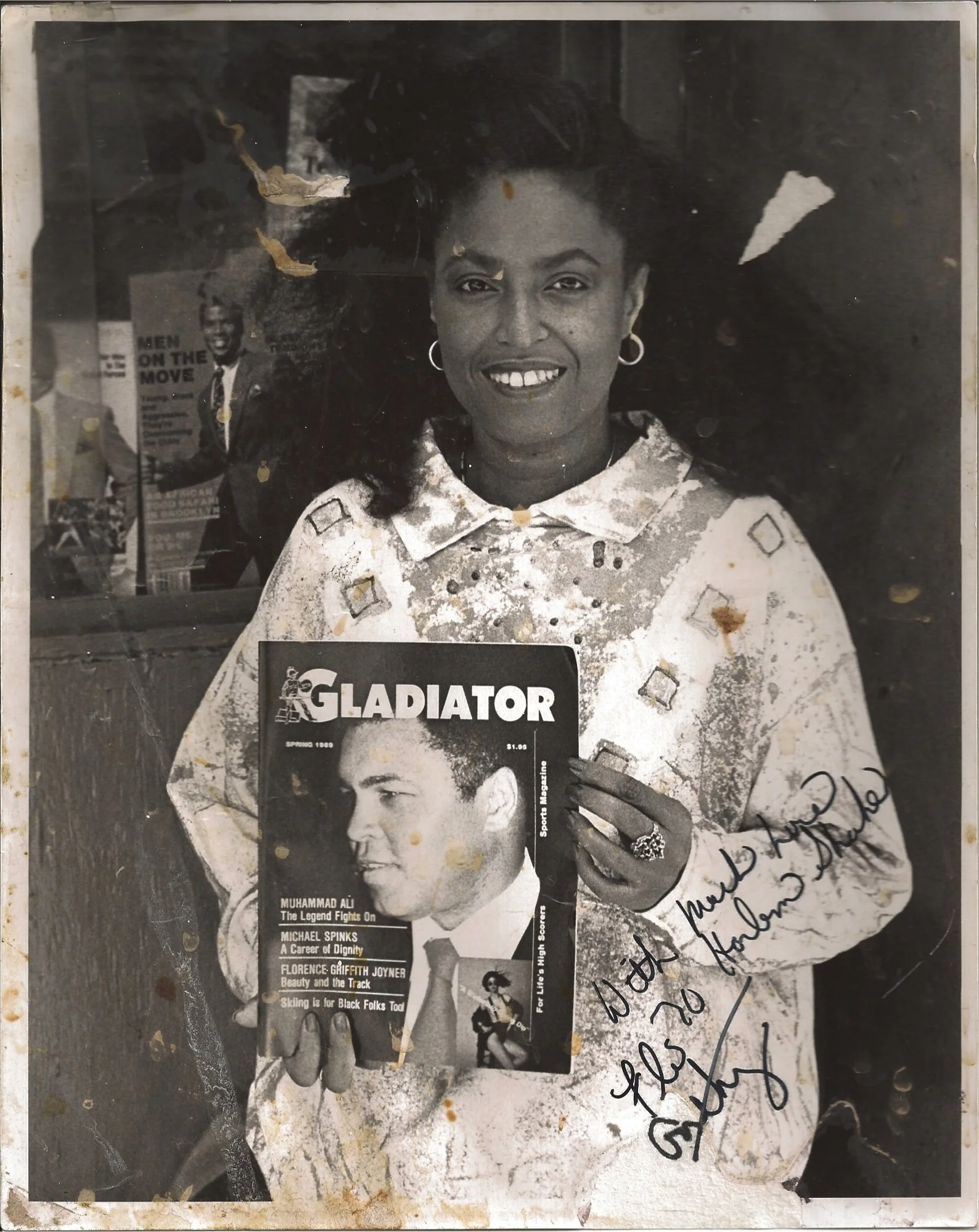 Black and white photograph of a smiling woman with curly hair holding a magazine featuring Muhammad Ali and Michael Spinks. She is wearing earrings and a patterned jacket, with handwritten notes and signatures on the photo.