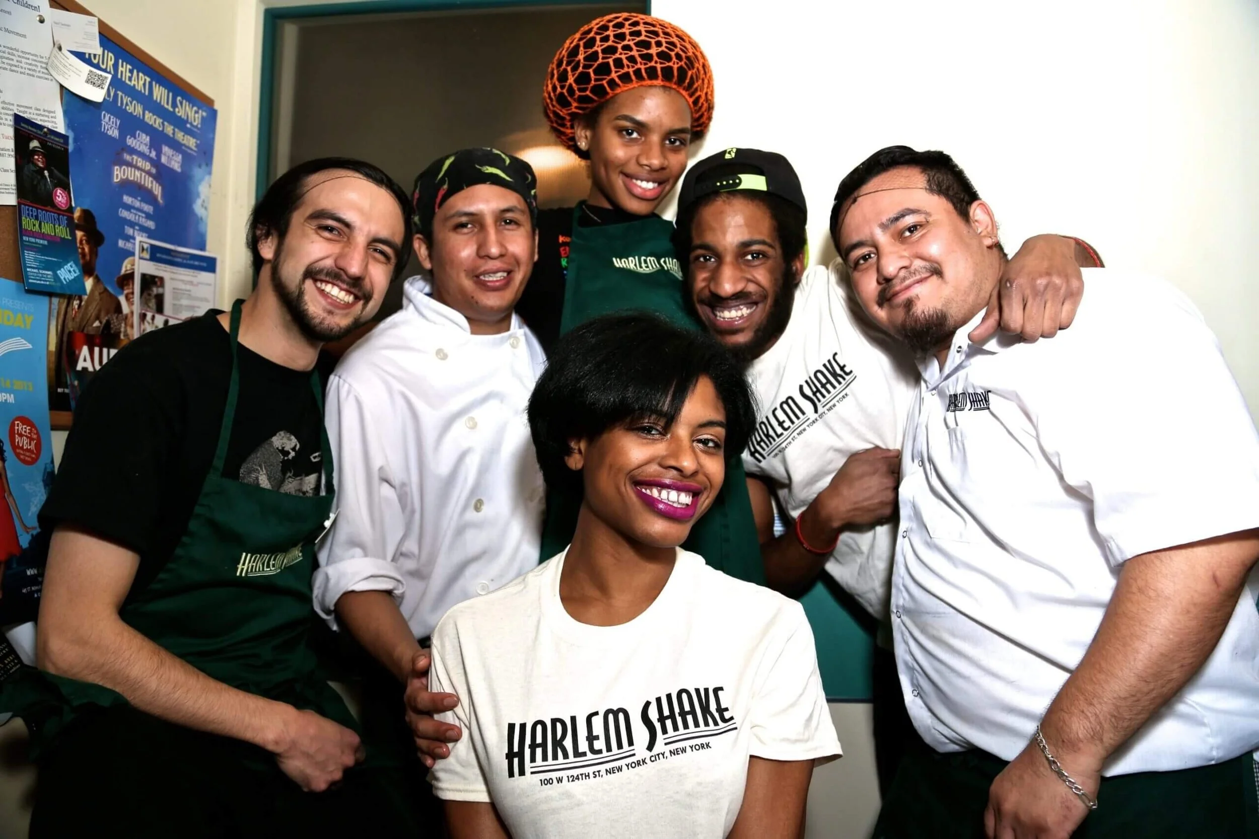 Group of six diverse people smiling and posing together inside a room, wearing casual clothing and some in chef uniforms. One woman in the center wears a bright orange head wrap and a green apron, with text that reads 'Harlem Shake, New York City, 100 W 124th St.' on her shirt. The background has posters and notes pinned on a corkboard.