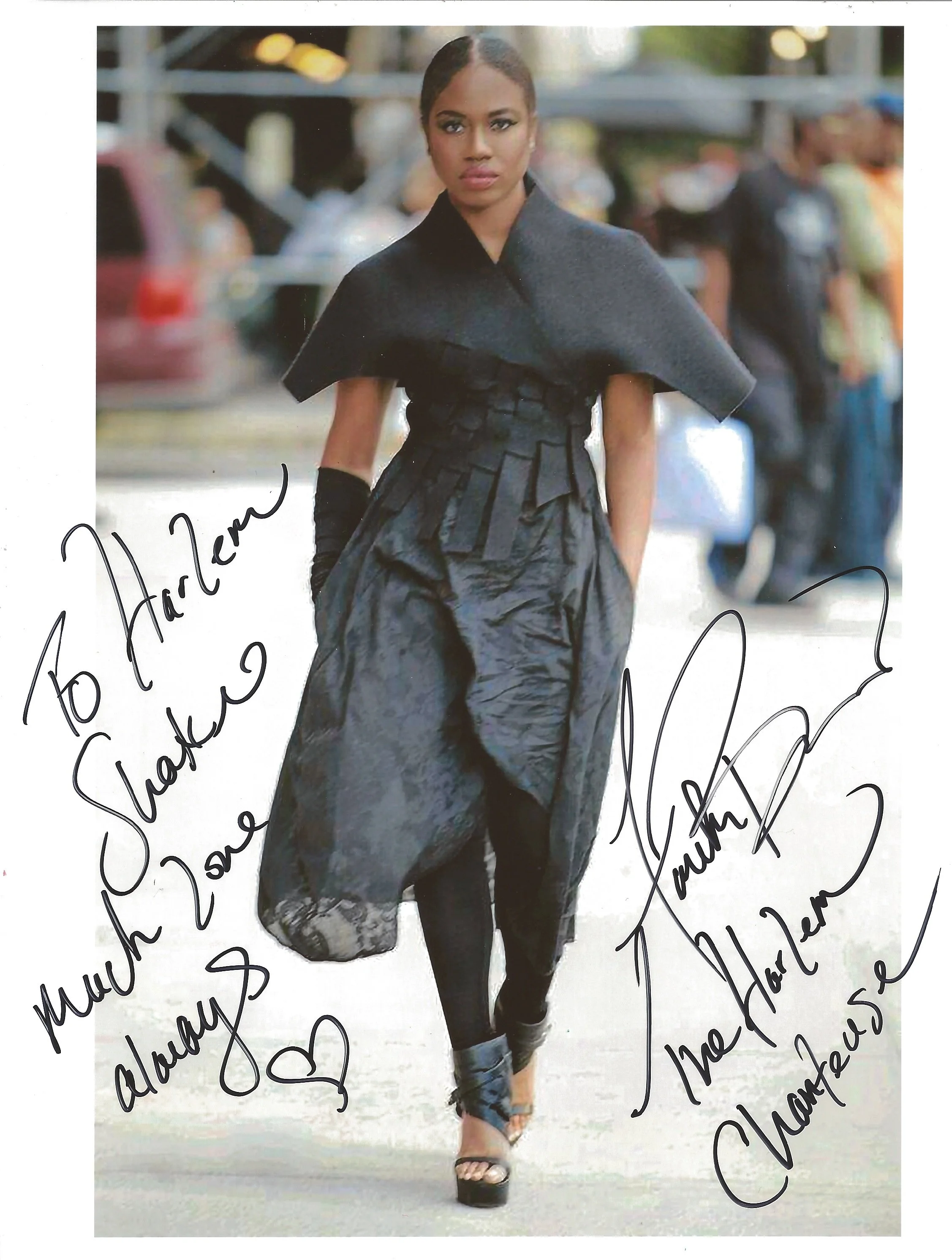 A woman with dark hair styled in finger waves, wearing a black dress, a gray wrap, and platform shoes, walking outdoors with a blurred background of people and structures. She has a serious expression and is looking directly at the camera. The image 