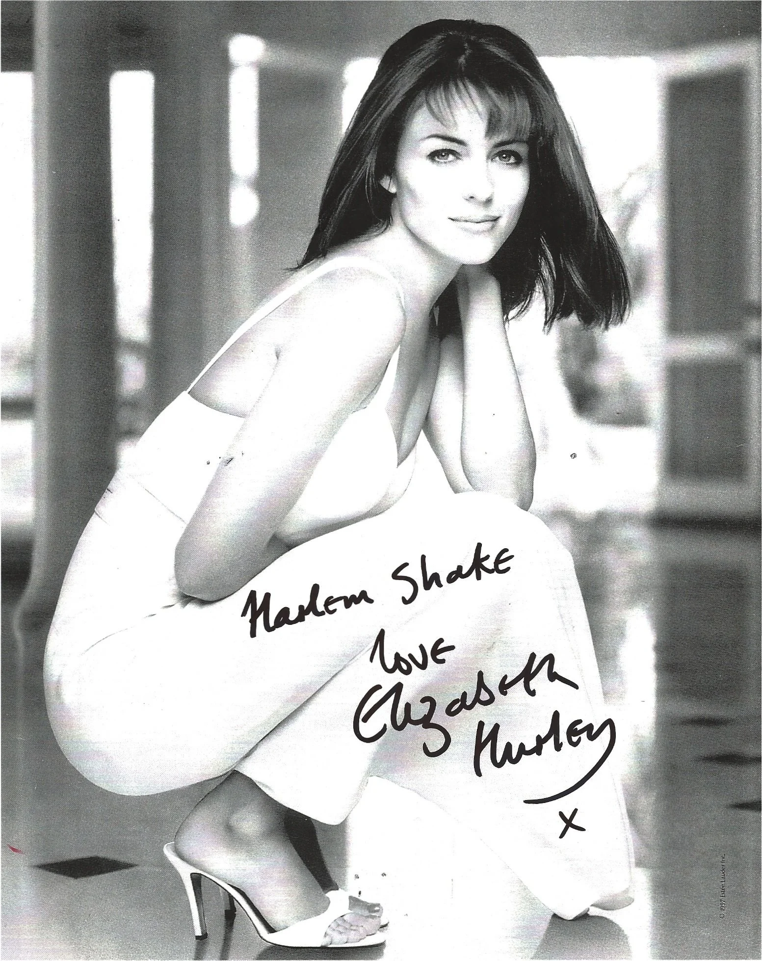 Black and white photograph of a woman with dark hair and light skin, sitting on a high heel shoe, wearing a dress with thin straps, smiling softly at the camera, with handwritten text on her dress that reads "Modern Shake Love Cuzedn & Wutney". The b