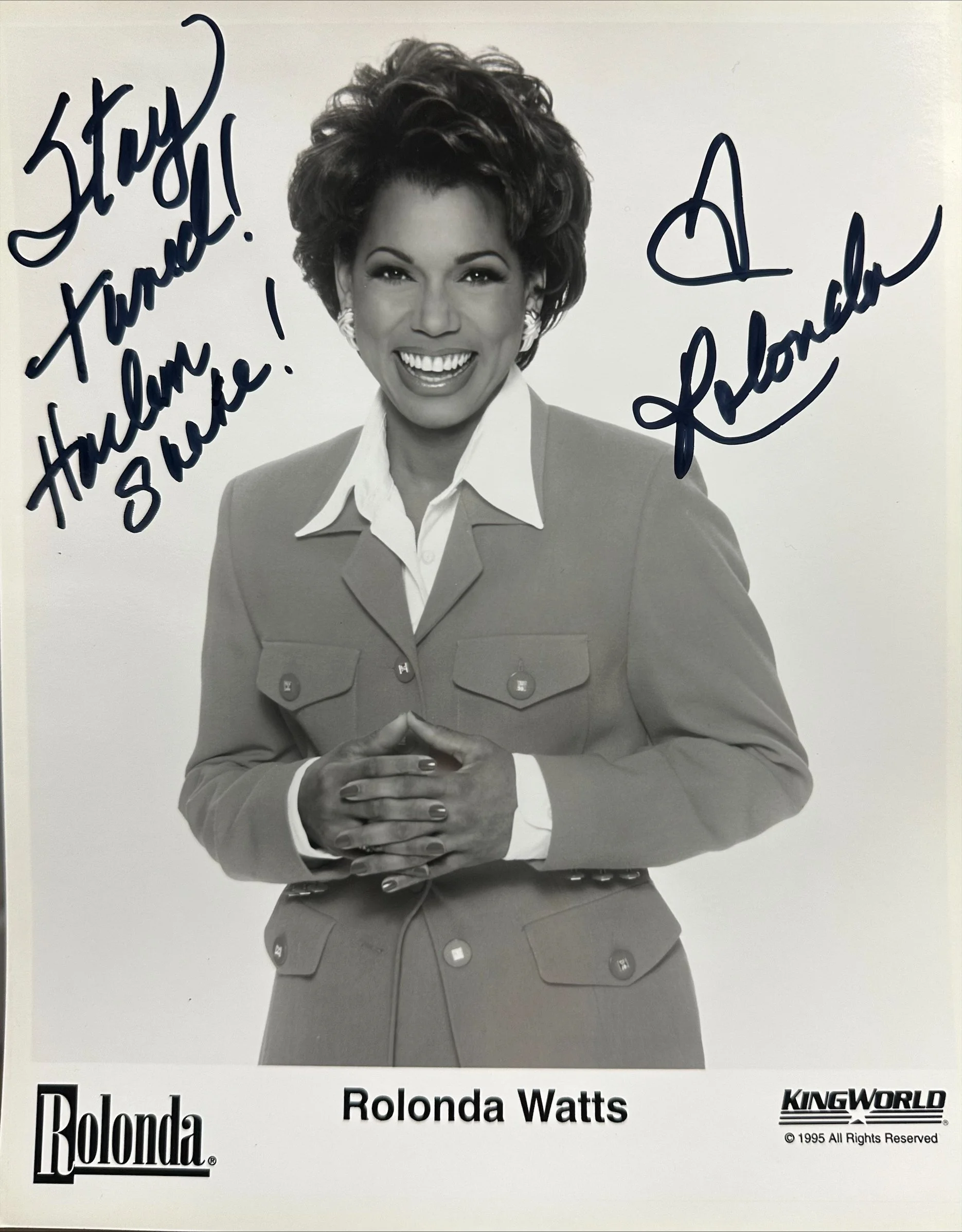 A black and white photo of a woman with short, curly hair smiling, wearing a suit jacket and white shirt, with her hands clasped at her waist. The photo has a handwritten message and signature in blue ink, with the text "Thank you! Thank you!" and a 