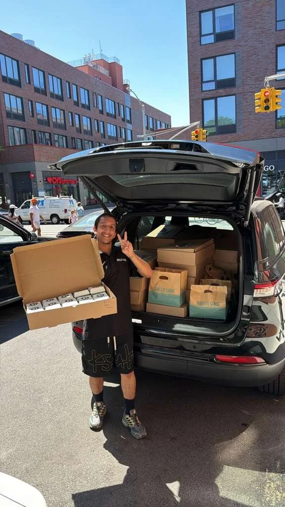 A man stands outside in front of an open SUV trunk filled with boxes and holds a large box with smaller boxes inside. The man is smiling and making a peace sign. The background shows city buildings and traffic lights.