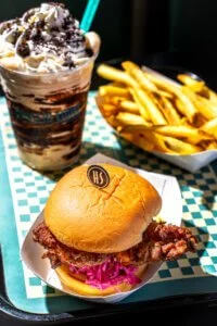 Chocolate milkshake topped with whipped cream and chocolate syrup, side of French fries, and a cheeseburger with lettuce and tomato in a bun