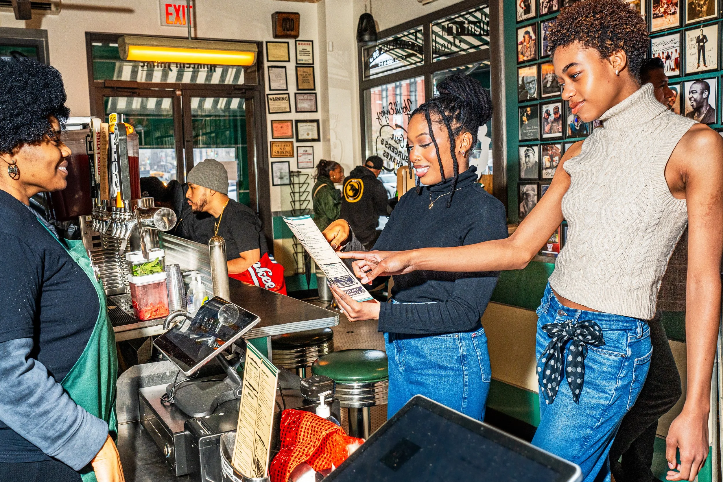 A group of young women at a restaurant counter, ordering food. One woman is reading the menu, while the server is smiling and wearing a green apron. In the background, other customers are sitting, and there are framed pictures on the walls.