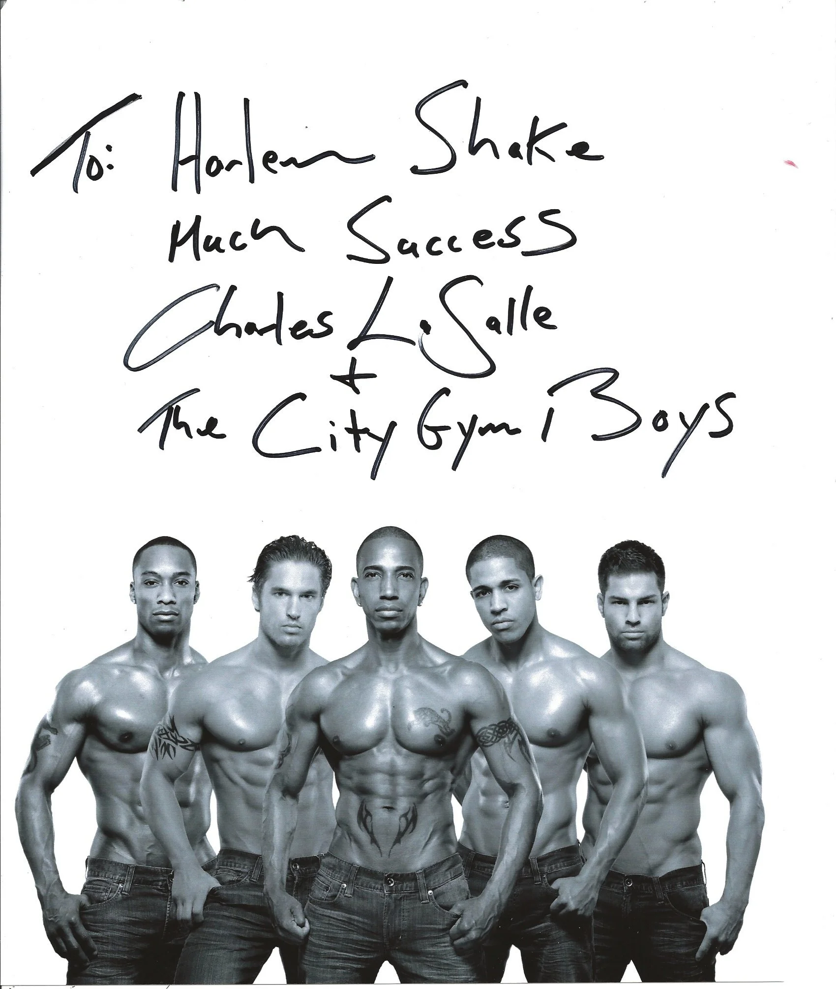 Black and white photo of six shirtless muscular men with tattoos standing in a row. They are wearing jeans and are looking at the camera.