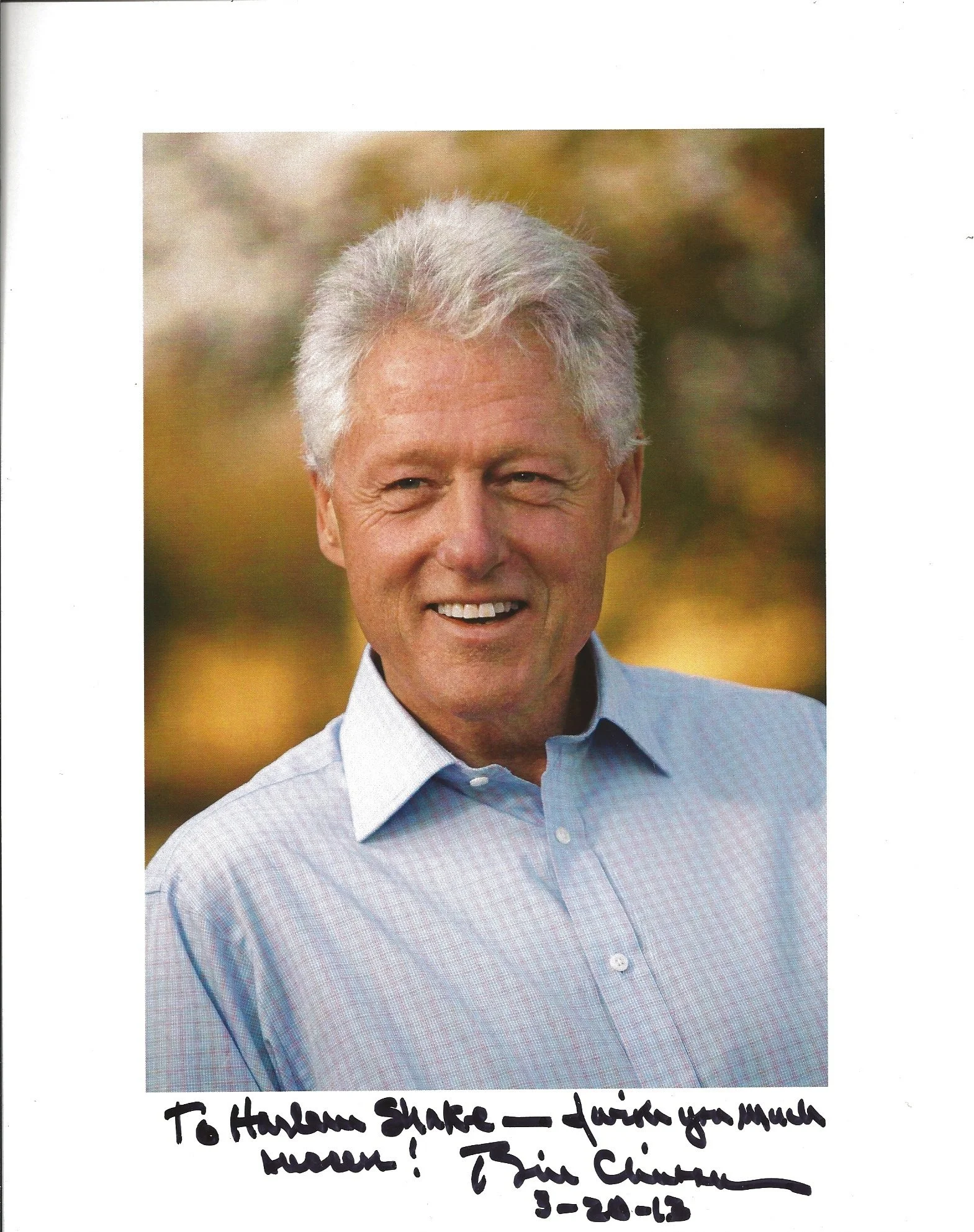 Photograph of a smiling older man with gray hair, outdoor background with blurred trees, wearing a light blue checkered shirt.