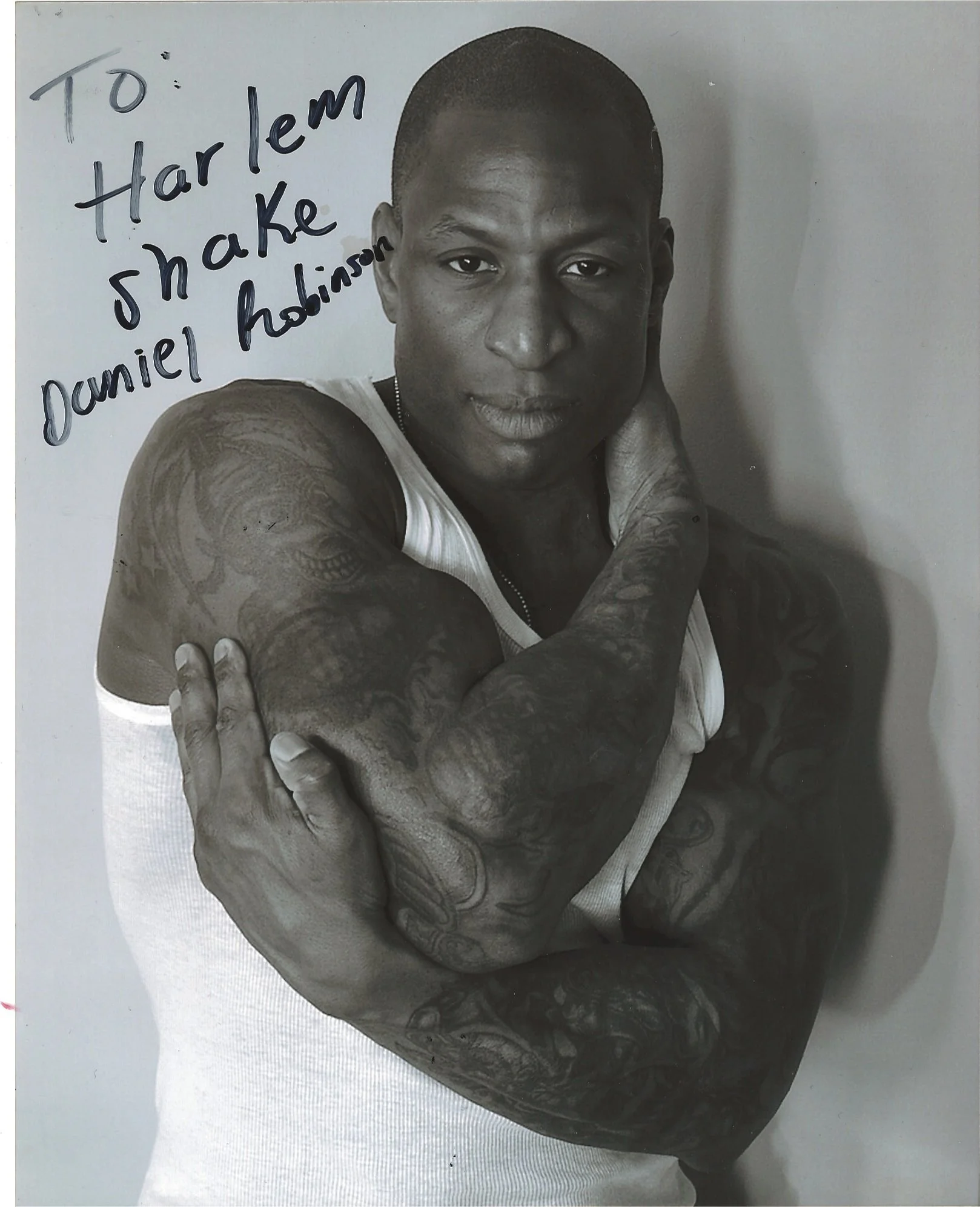 A black and white photograph of a young man with tattoos on his arms, wearing a sleeveless shirt, looking directly into the camera with a serious expression. Handwritten text on the top left corner reads, "To Harlem shake, Daniel Robinson."