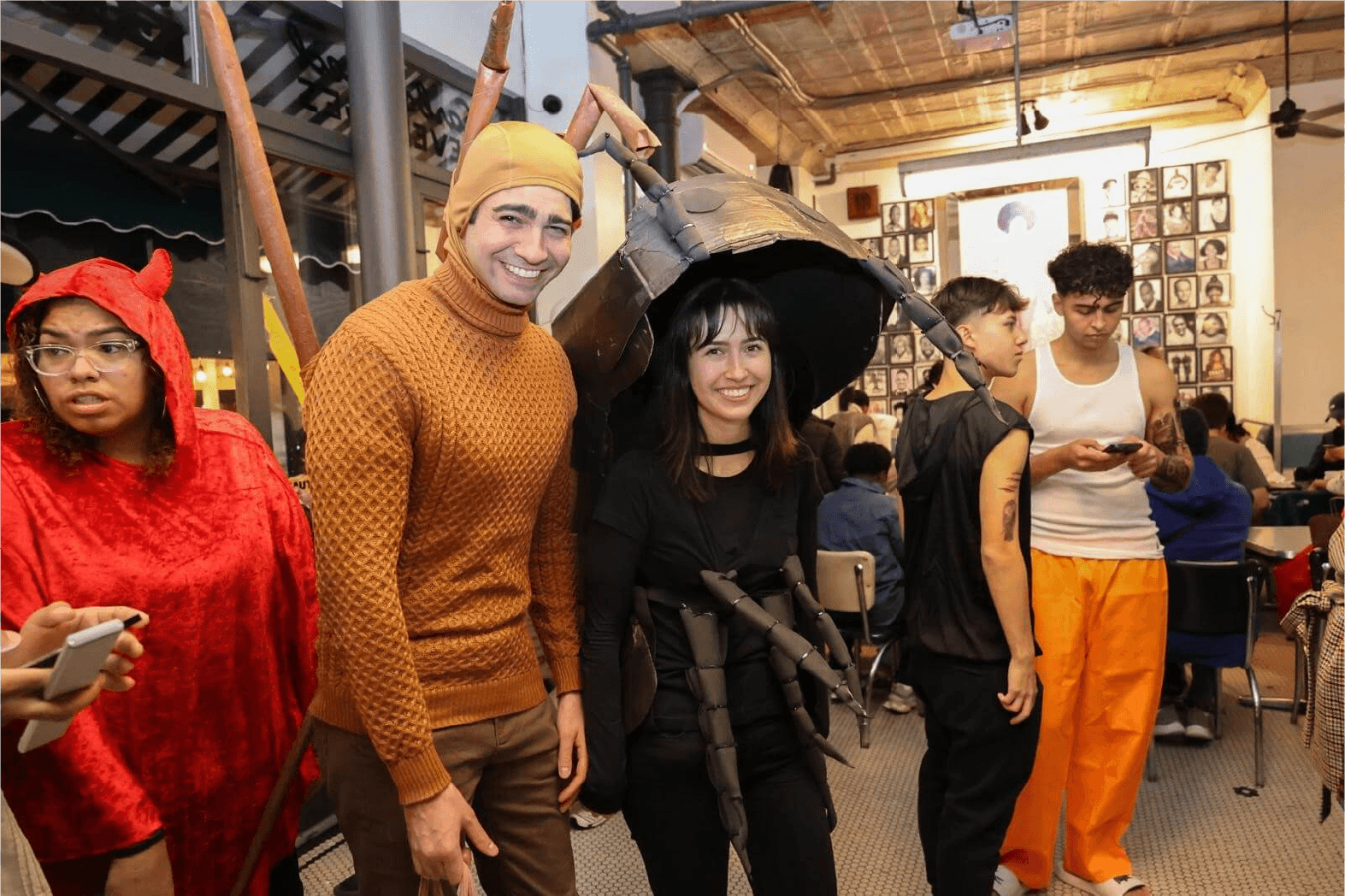 Group of five people dressed in costumes at a social gathering, posing for photo in a decorated indoor space, with people sitting at tables in the background.