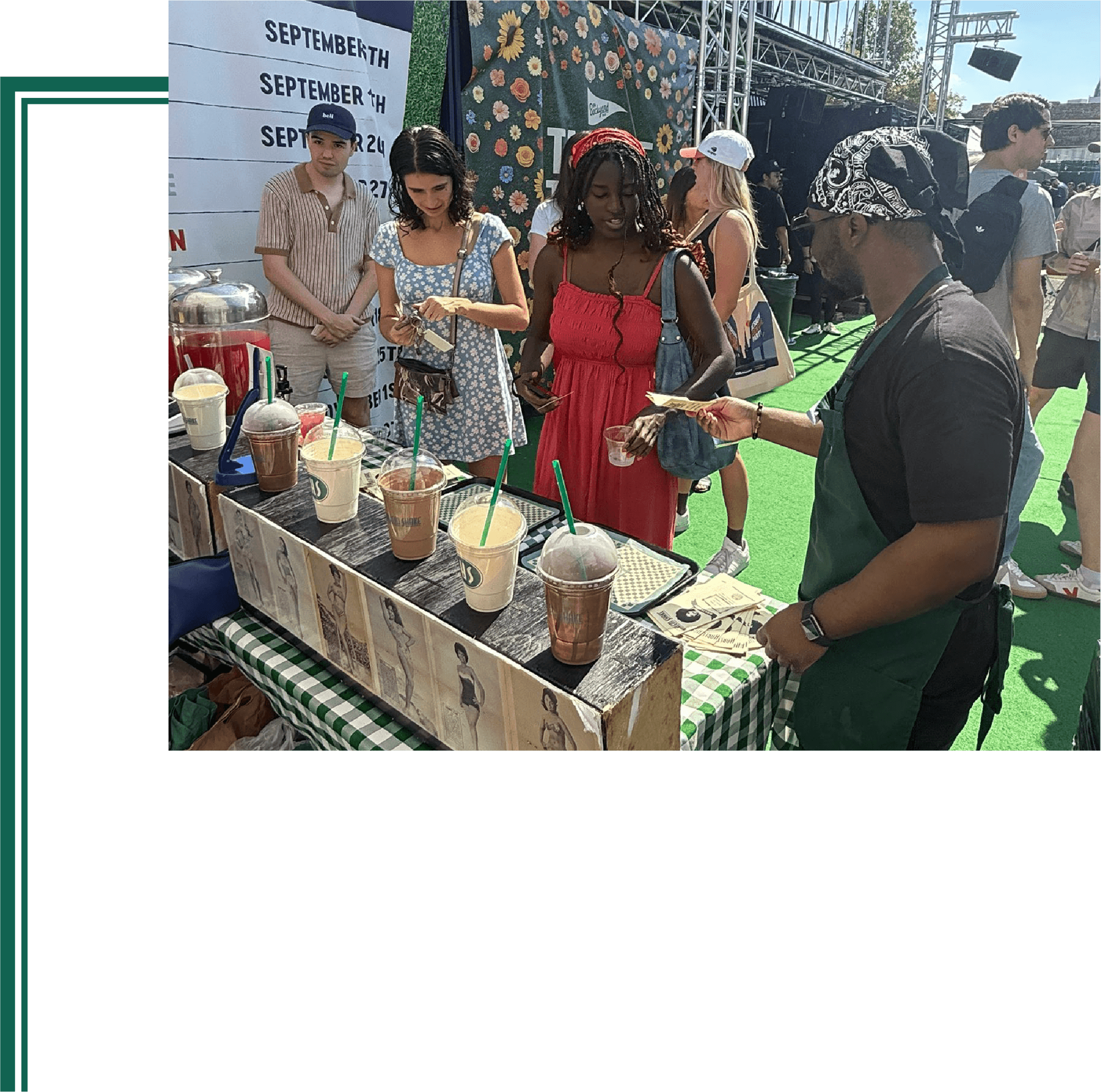 A group of people at an outdoor event near a booth with drinks. People are standing and looking at a menu or flyer, with a large floral banner in the background and a sunny sky above.