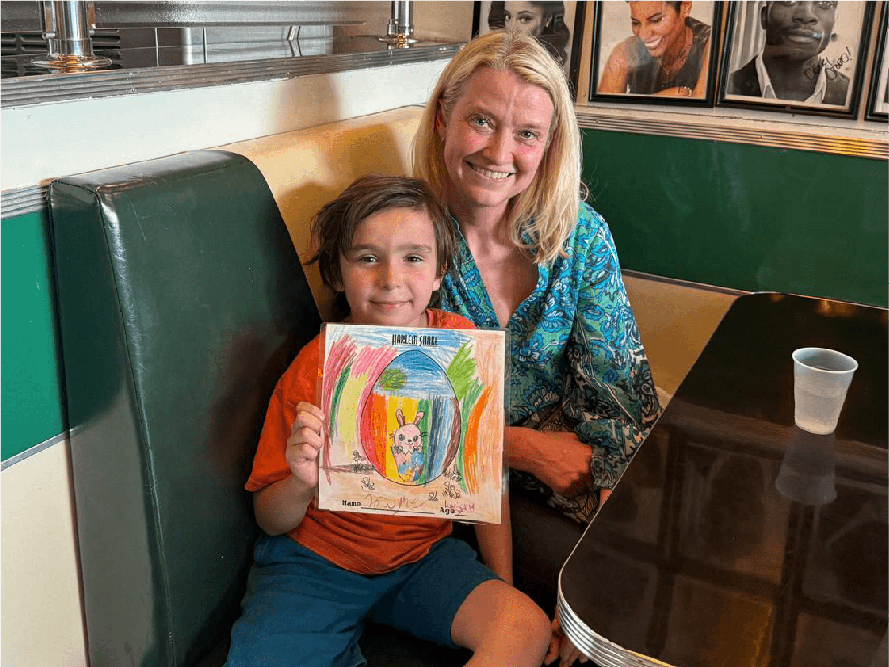A young boy and a woman sitting in a booth at a restaurant. The boy is holding a colorful drawing of a unicorn with a rainbow background. The woman is smiling, and there are framed black-and-white portraits on the wall behind them. The table has a plastic cup on it.