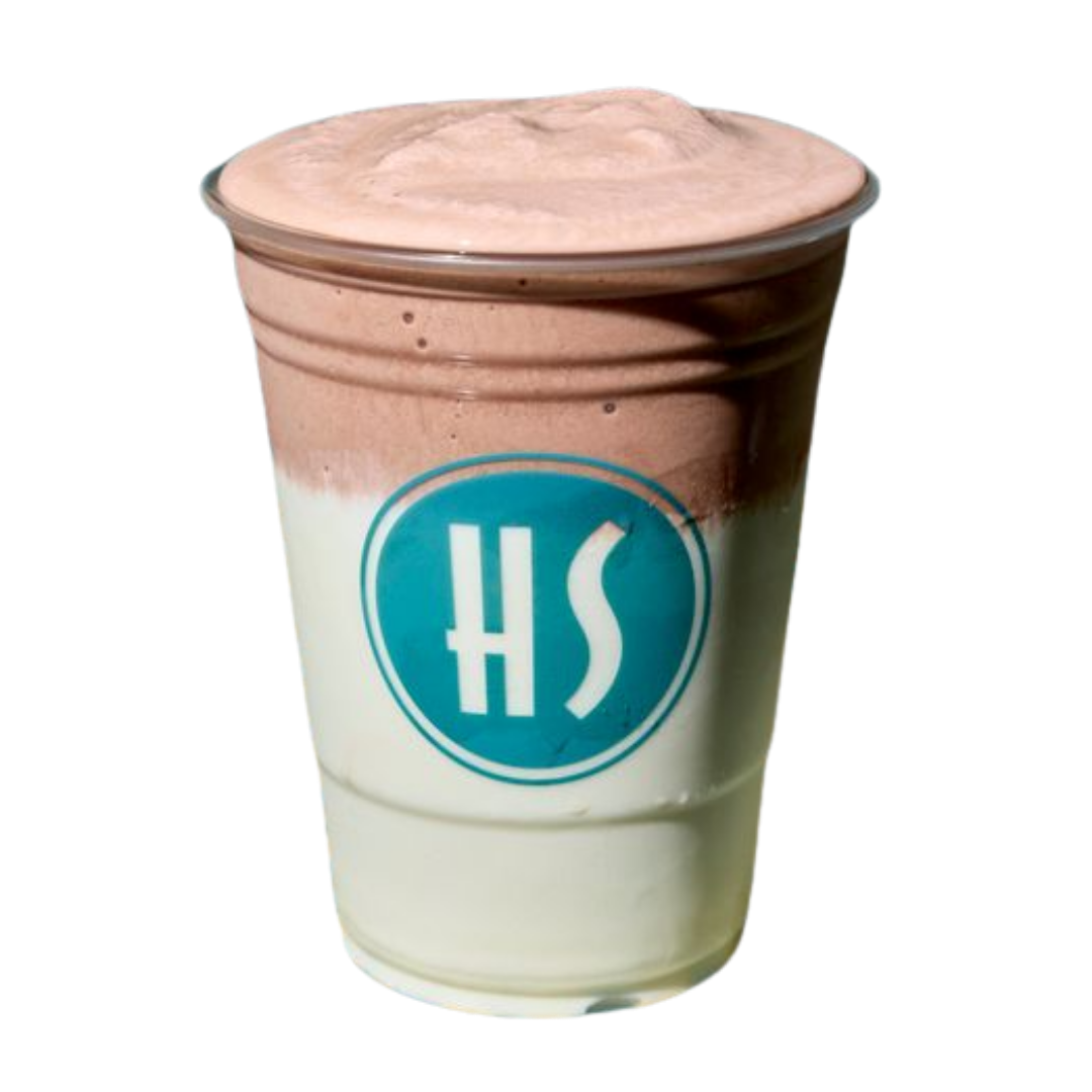 A clear plastic cup with two layered beverages, one white and one chocolate-colored, topped with a frothy foam, with a logo that has the letters 'HS' in a blue circle.