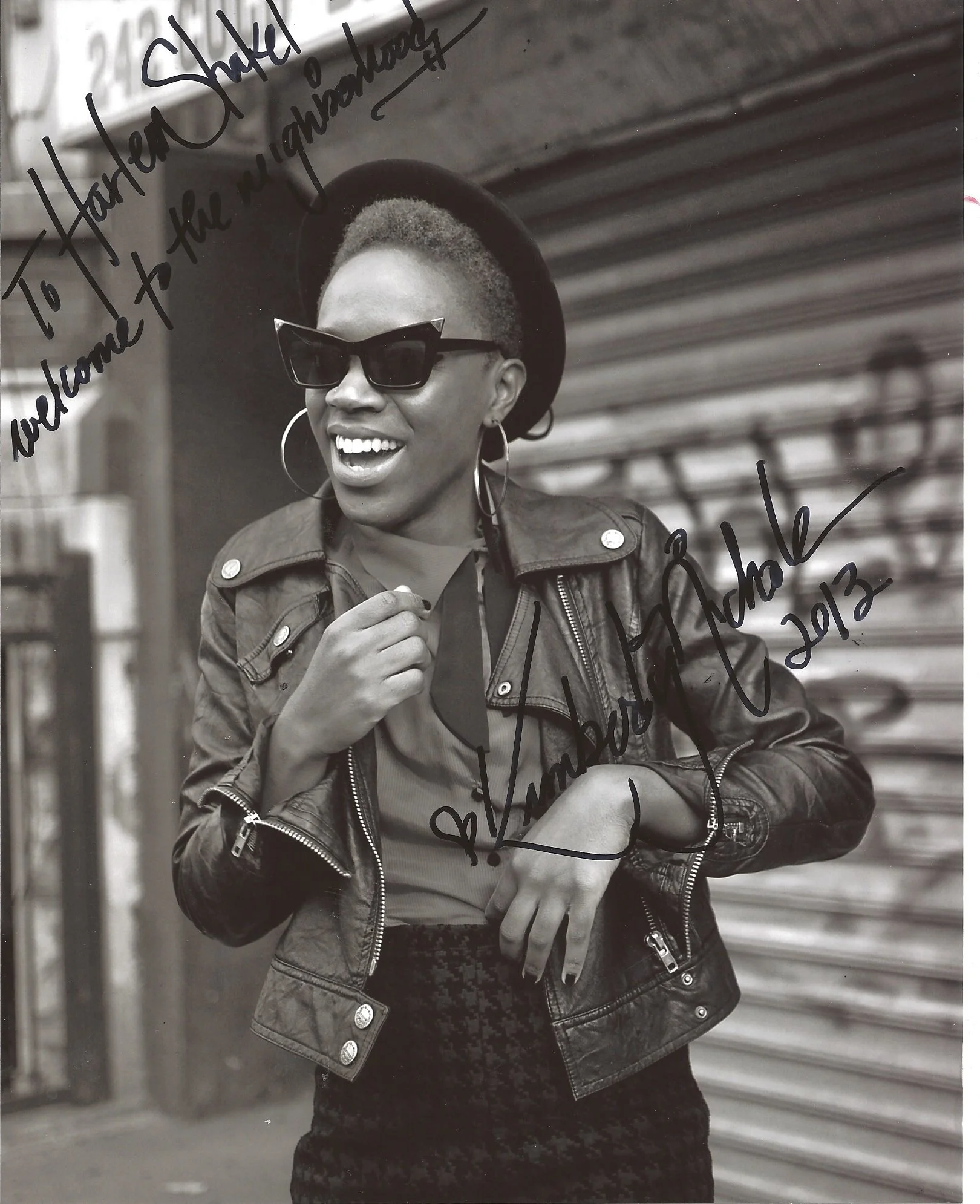 A woman wearing sunglasses, a hat, a leather jacket, and hoop earrings, smiling with her teeth showing, standing in front of a metal background.