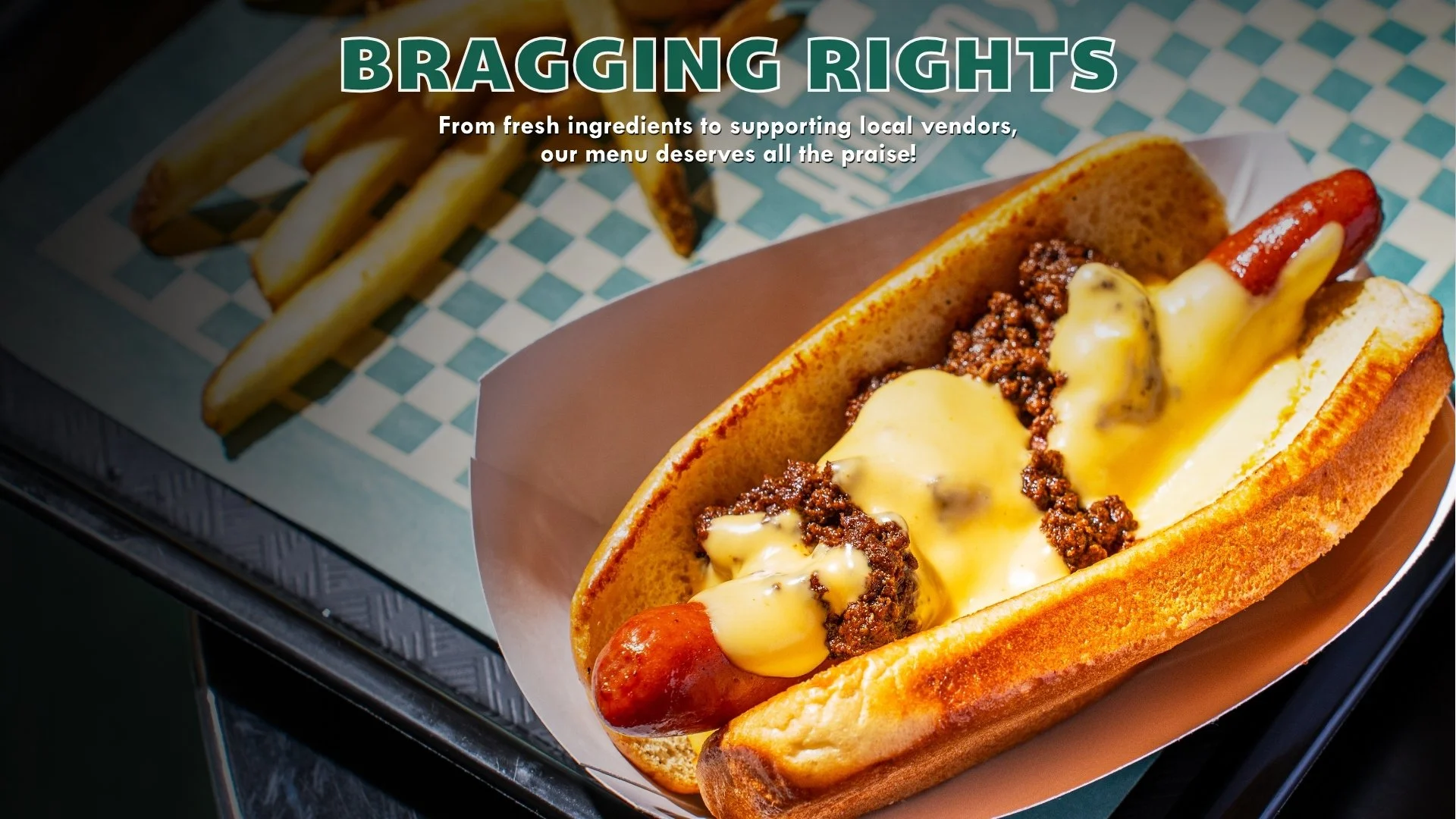A hot dog in a bun topped with melted cheese, cooked ground beef, and a sausage, served on a pink paper boat with fries in the background.