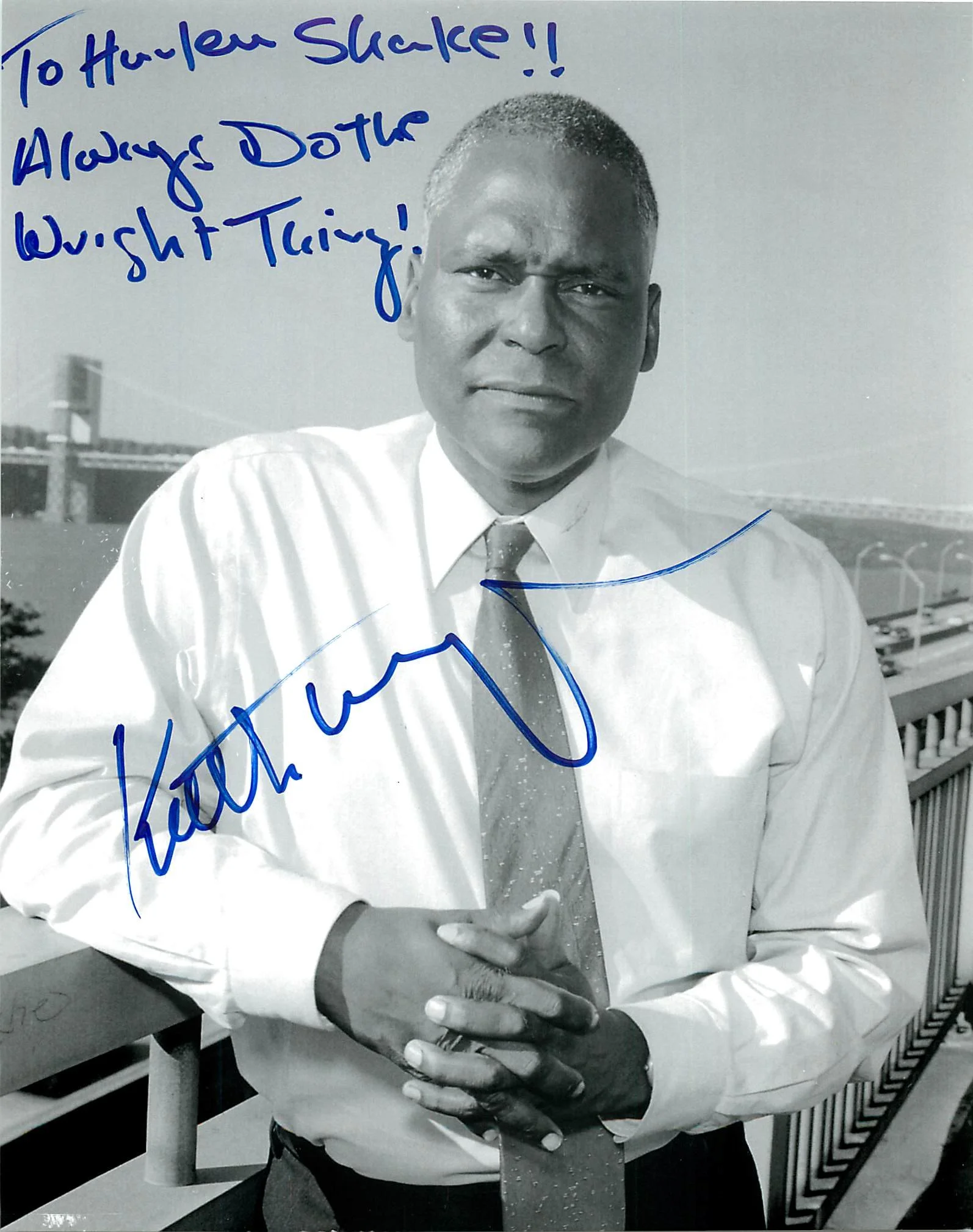 A black-and-white photograph of a man dressed in a white shirt and tie, standing outdoors near a bridge or railing. He has short hair and is looking at the camera with a serious expression. Handwritten blue ink notes overlay the image, including a si