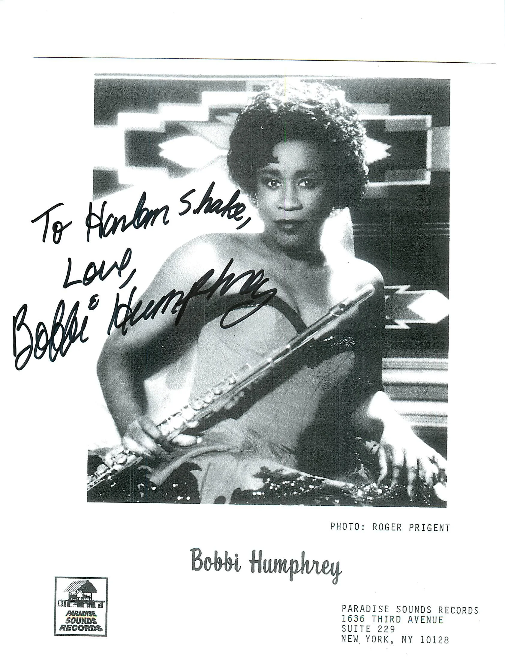 A black-and-white promotional flyer featuring a photograph of a woman with short curly hair, holding a flute and looking directly at the camera. The flyer contains handwritten text that reads, 'To Hannah Shake, Love' followed by a signature. The flye