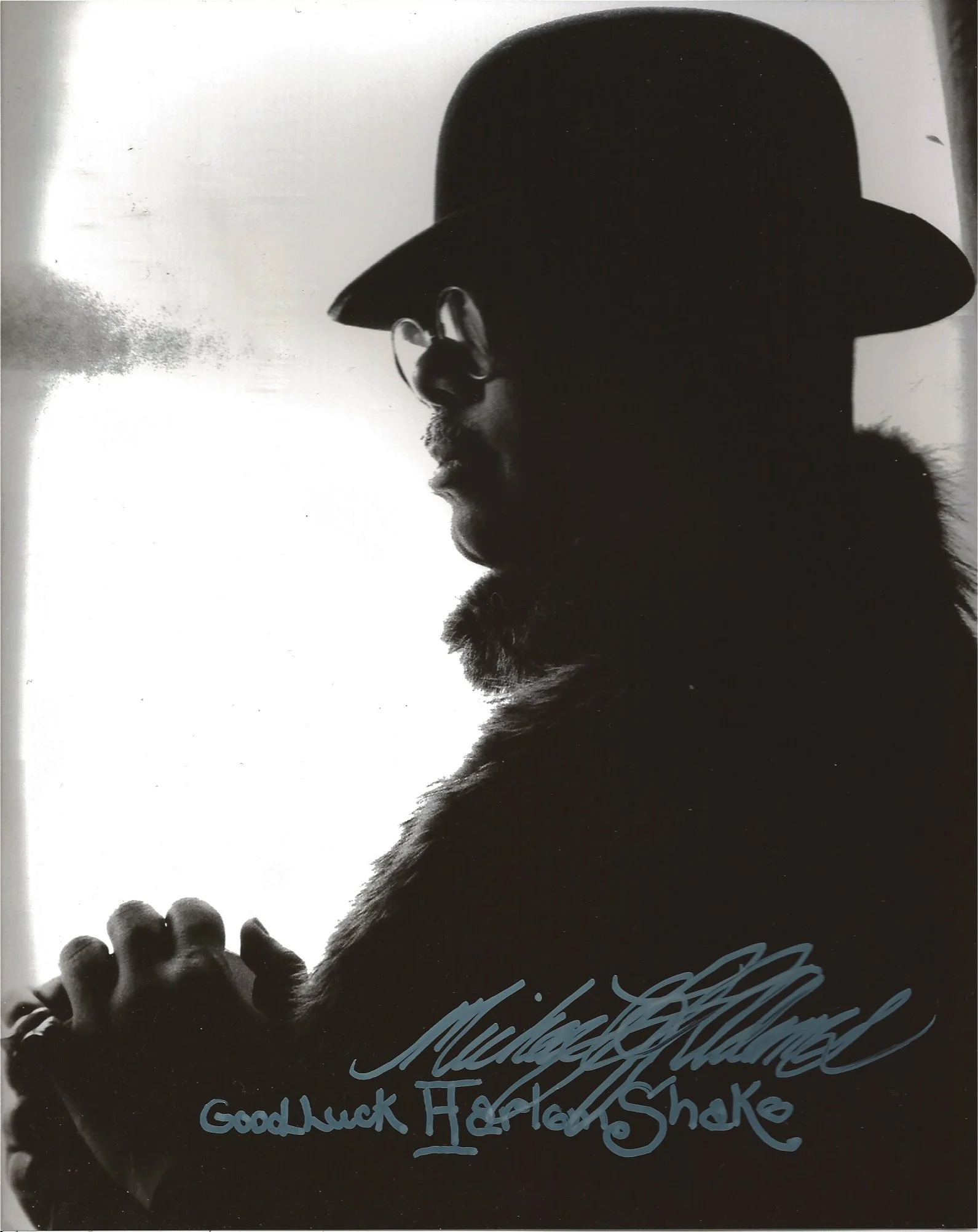 Black-and-white image of a person in profile wearing a wide-brim hat and glasses, with a talk bubble saying 'Good Luck Harlem Shake', and a handwritten signature below.