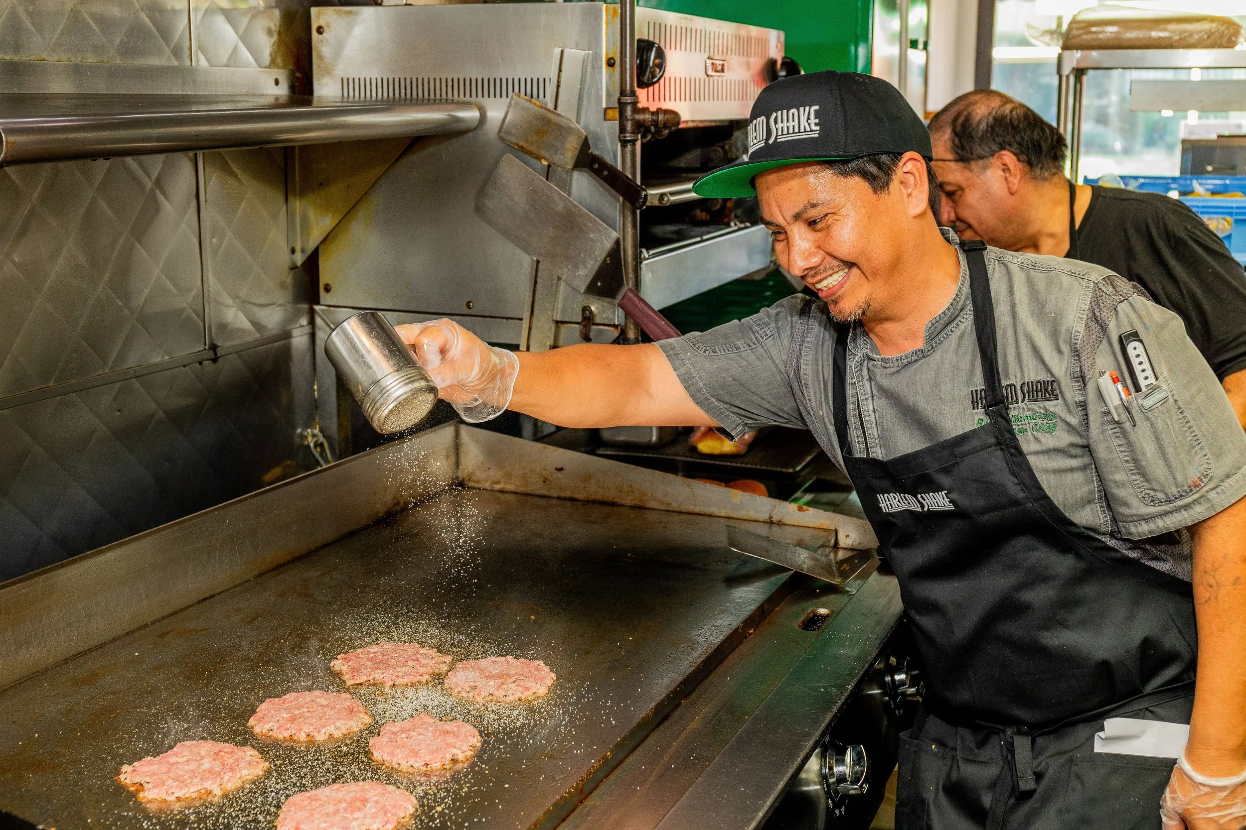 A man smiling while seasoning raw hamburger patties on a griddle in a kitchen, with another person working nearby in the background.