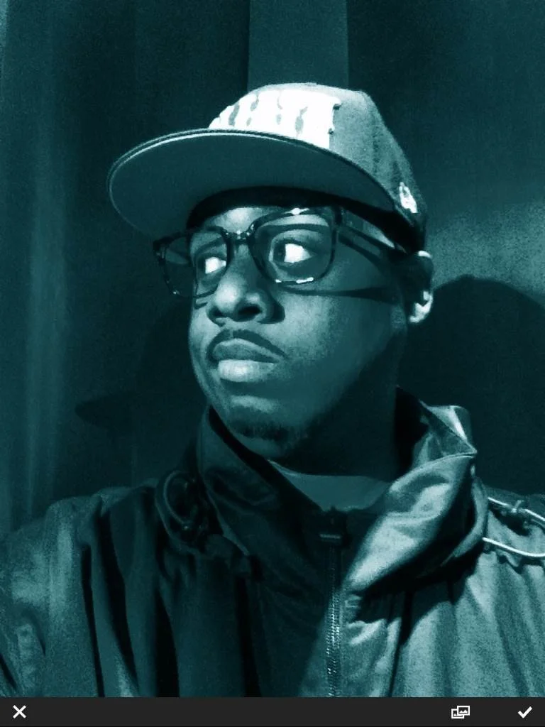 A young man wearing glasses, a baseball cap, and a dark jacket, looking to his left with a neutral expression.