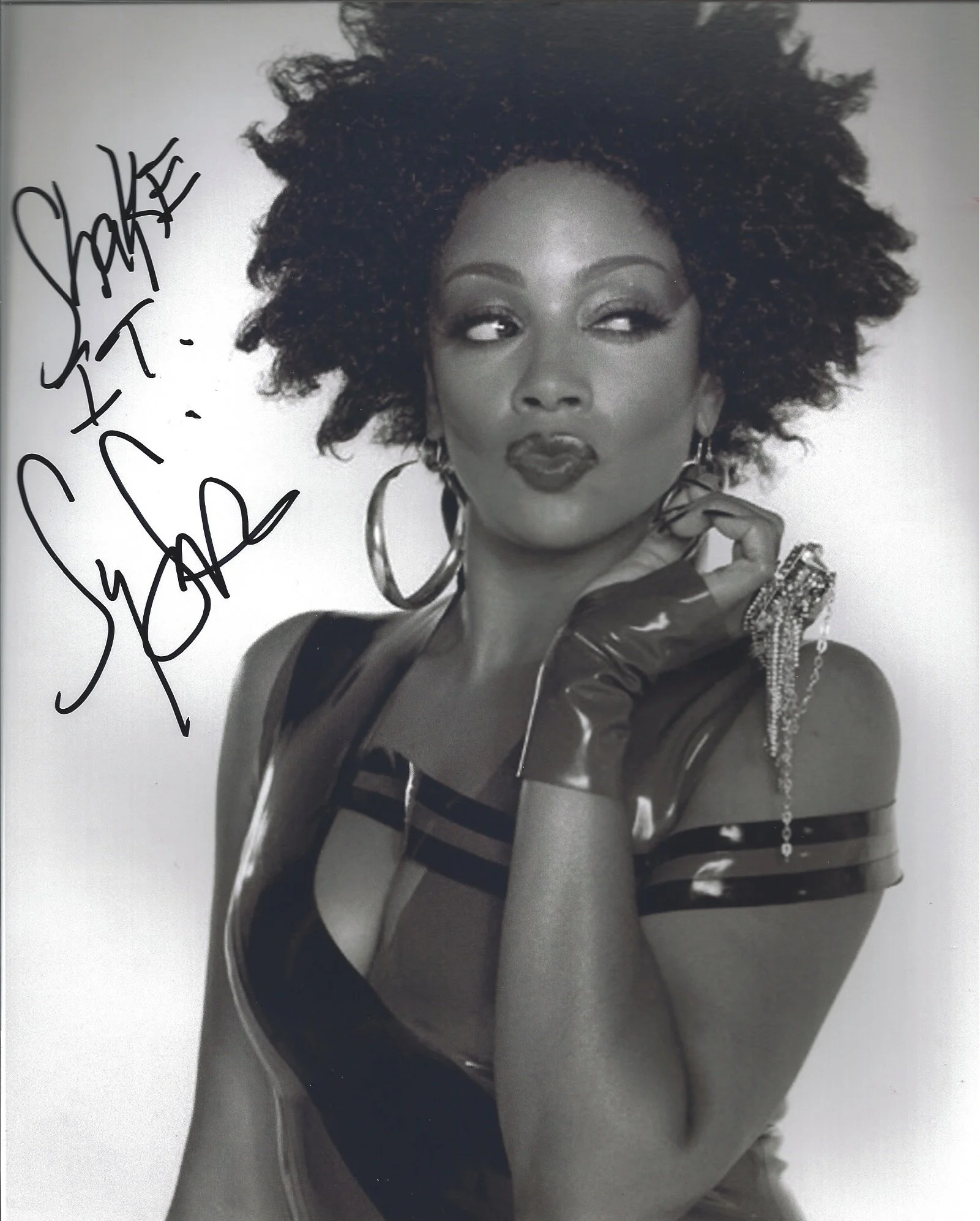 Black-and-white photograph of a woman with voluminous, curly hair making a playful facial expression with her eyes looking to the side and lips pursed. She is wearing large hoop earrings, rings, a striped top, and gloves, with a chain hanging from he