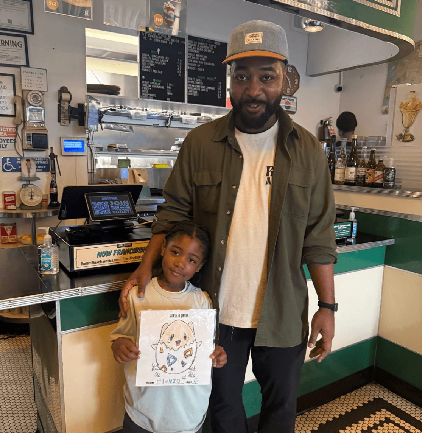 A man and a young girl standing inside a restaurant near the counter. The girl is holding a drawing of an animal character labeled 'Harlem Shake' with a name and age written on it. The man has a beard, wears a gray cap, and a green jacket. The background shows menu boards, bottles, and a cash register.