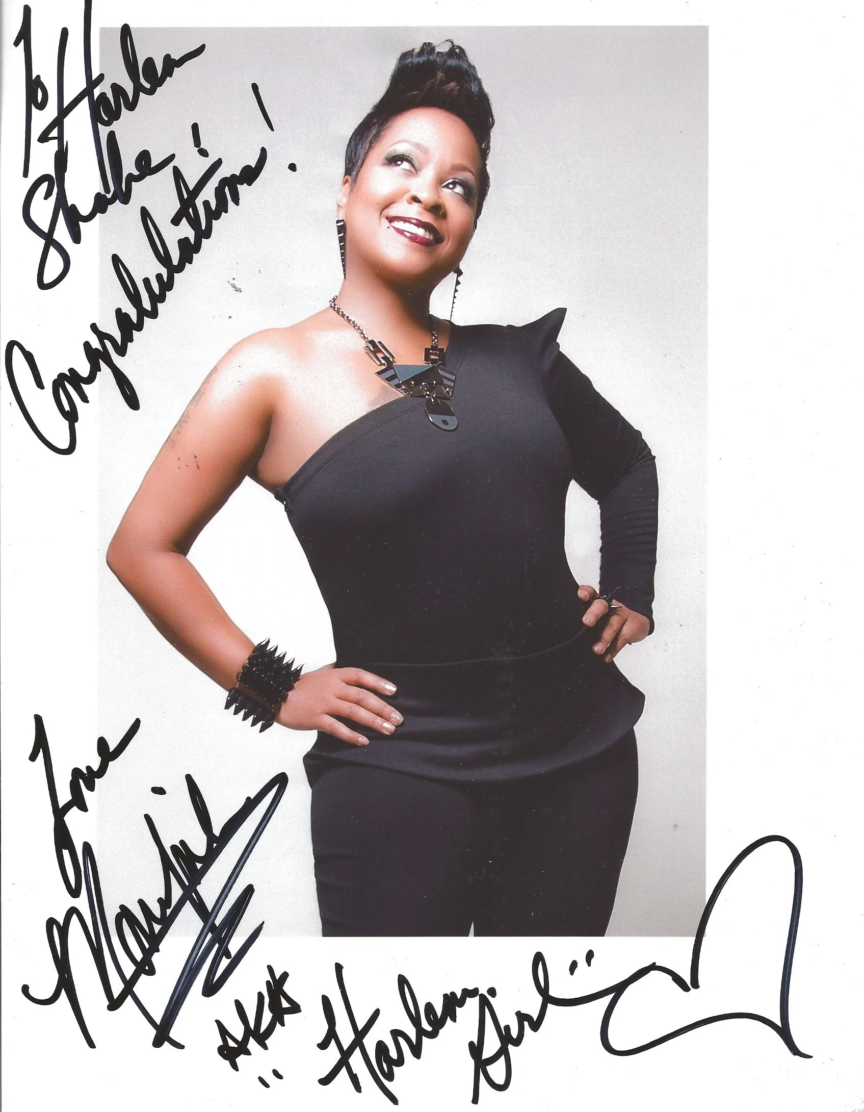 Photograph of a woman with short dark hair, wearing a black, off-the-shoulder dress, and jewelry, standing against a plain white background. She is smiling and looking upwards. The picture is autographed with a handwritten message.