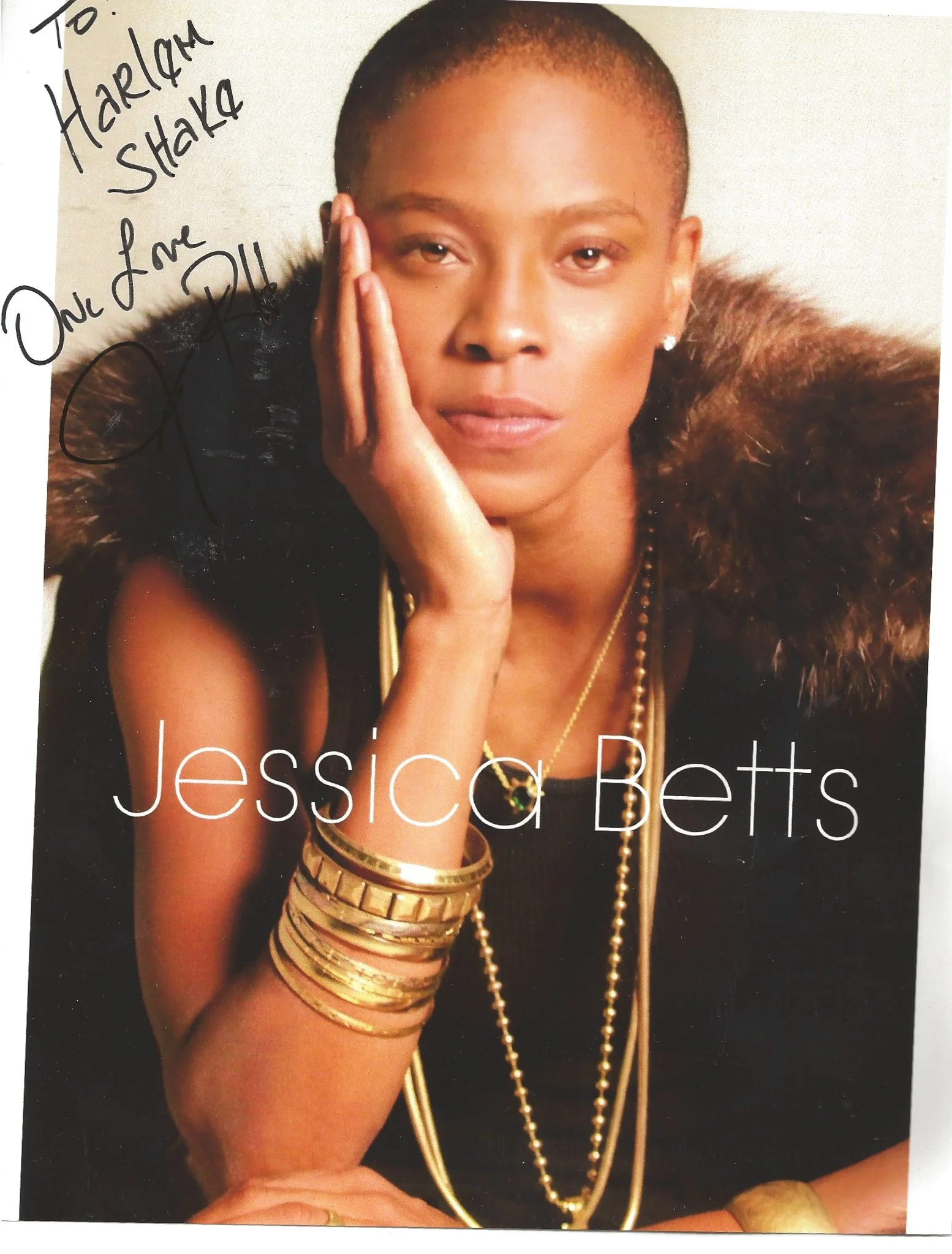 A woman with short hair, wearing multiple gold bracelets, a gold necklace, and earrings, resting her face against her hand, looking at the camera with a neutral expression.