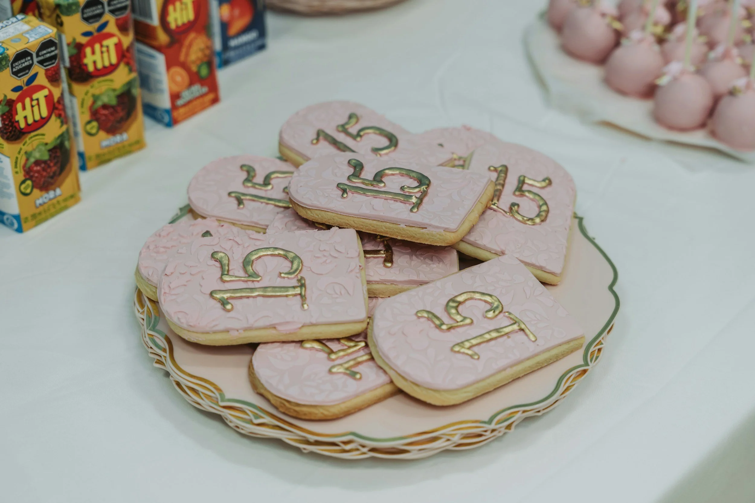A plate of pink cookies decorated with gold writing and designs, with the number 15 on them, surrounded by boxes of fruit juice and a tray of pink cake balls in the background.