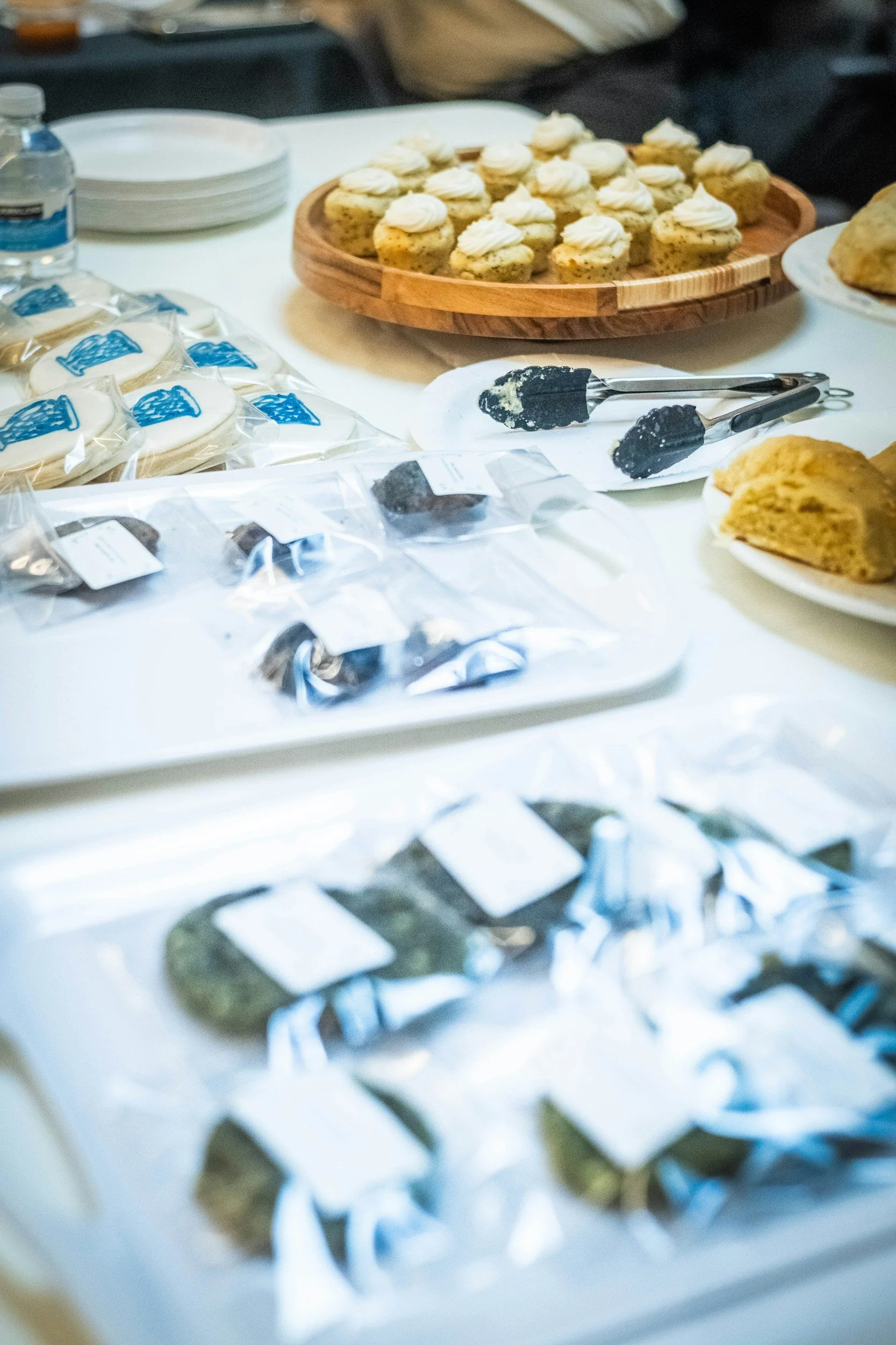 Assorted cookies and baked goods on a table, including cookies with white frosting, black decorated cookies, and other pastries, with some packaged and some on plates, in a casual setting.