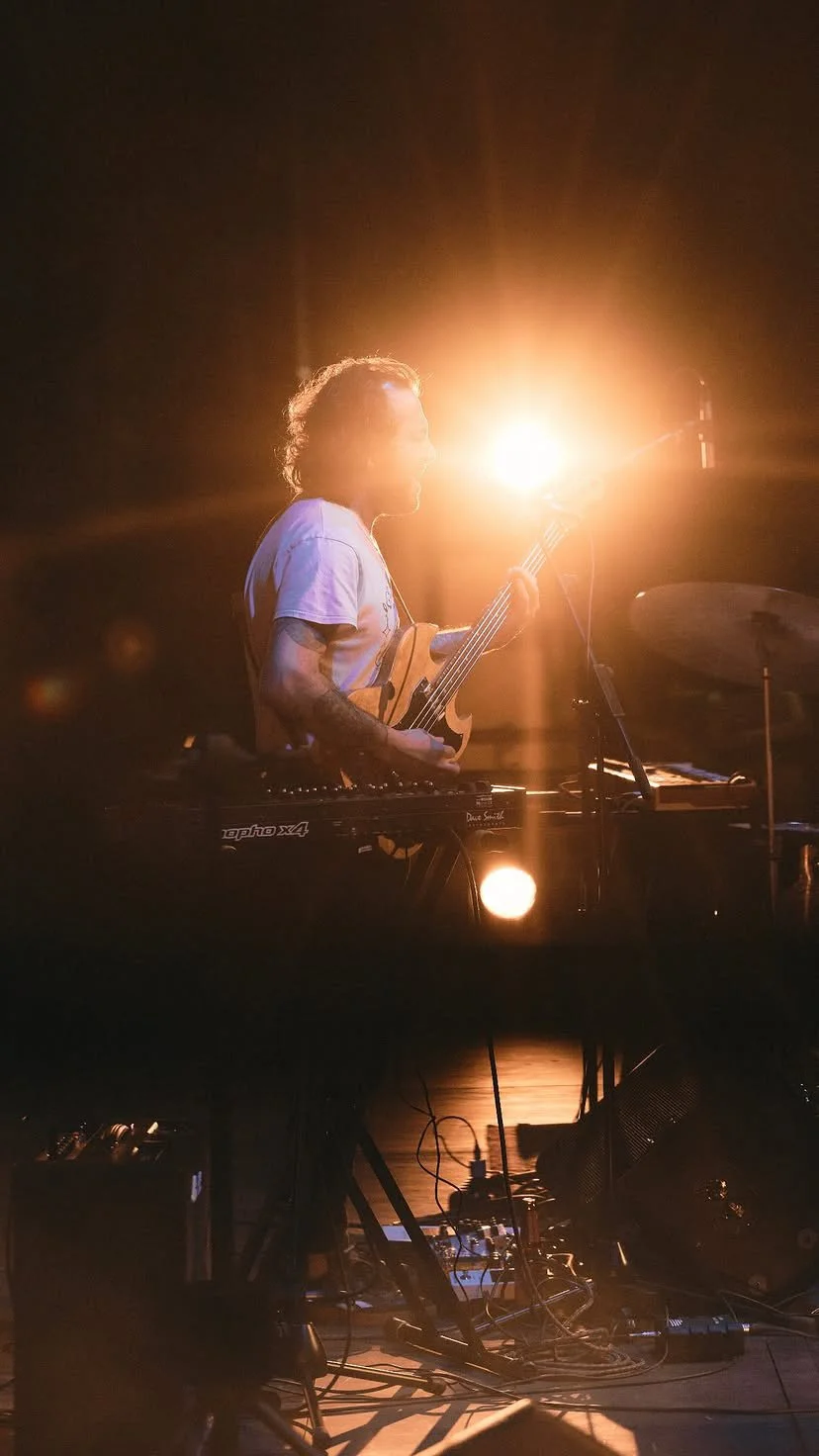 Un músico tocando guitarra y teclado en un escenario con iluminación brillante en la parte trasera.