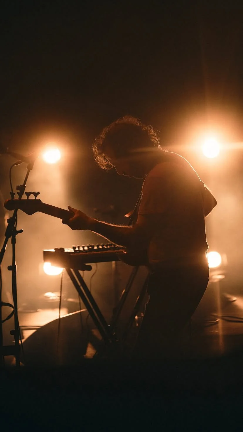 Persona tocando guitarra y teclado en un escenario con luces brillantes al fondo.