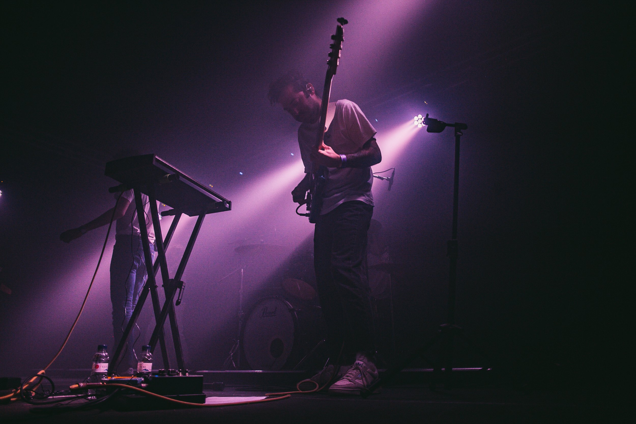 Un músico tocando la guitarra eléctrica en un escenario con iluminación púrpura, con un teclado y una botella de agua en el suelo.