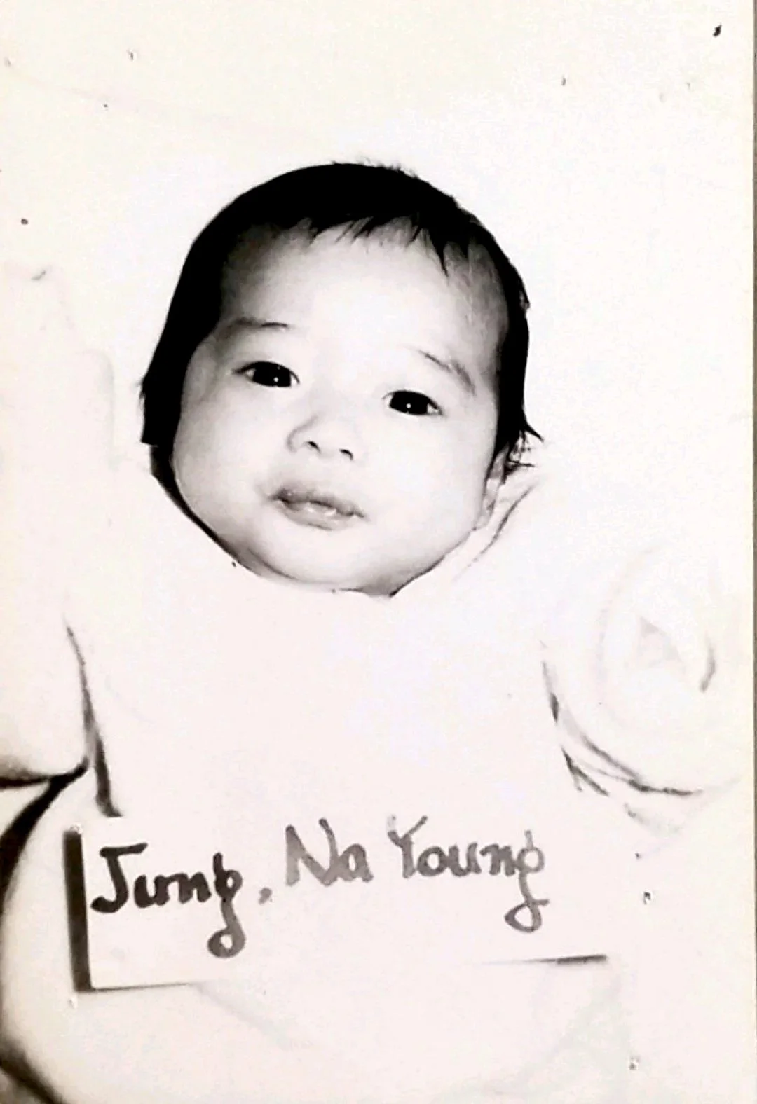 Black and white close-up of baby Nayoung Jung, lying on a soft surface. She has short, dark hair and dark eyes.