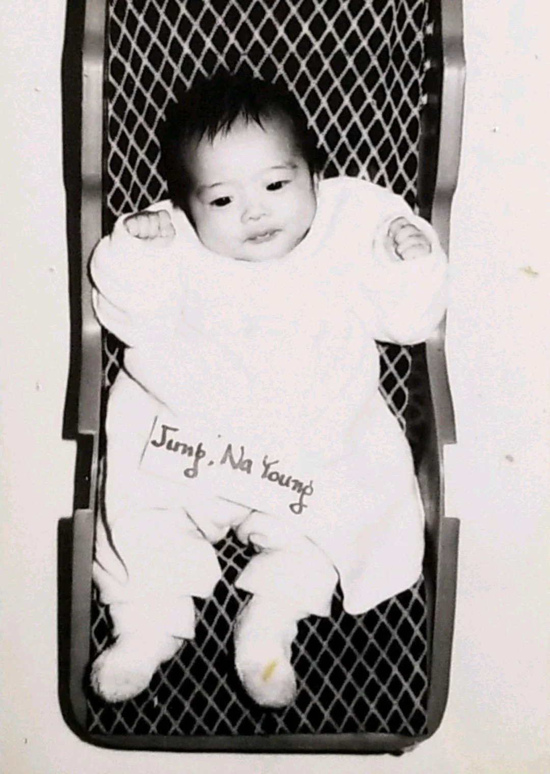 Black and white photo of a baby Nayoung Jung, strapped to a baby carrier and wearing a light-colored outfit while looking at the camera.