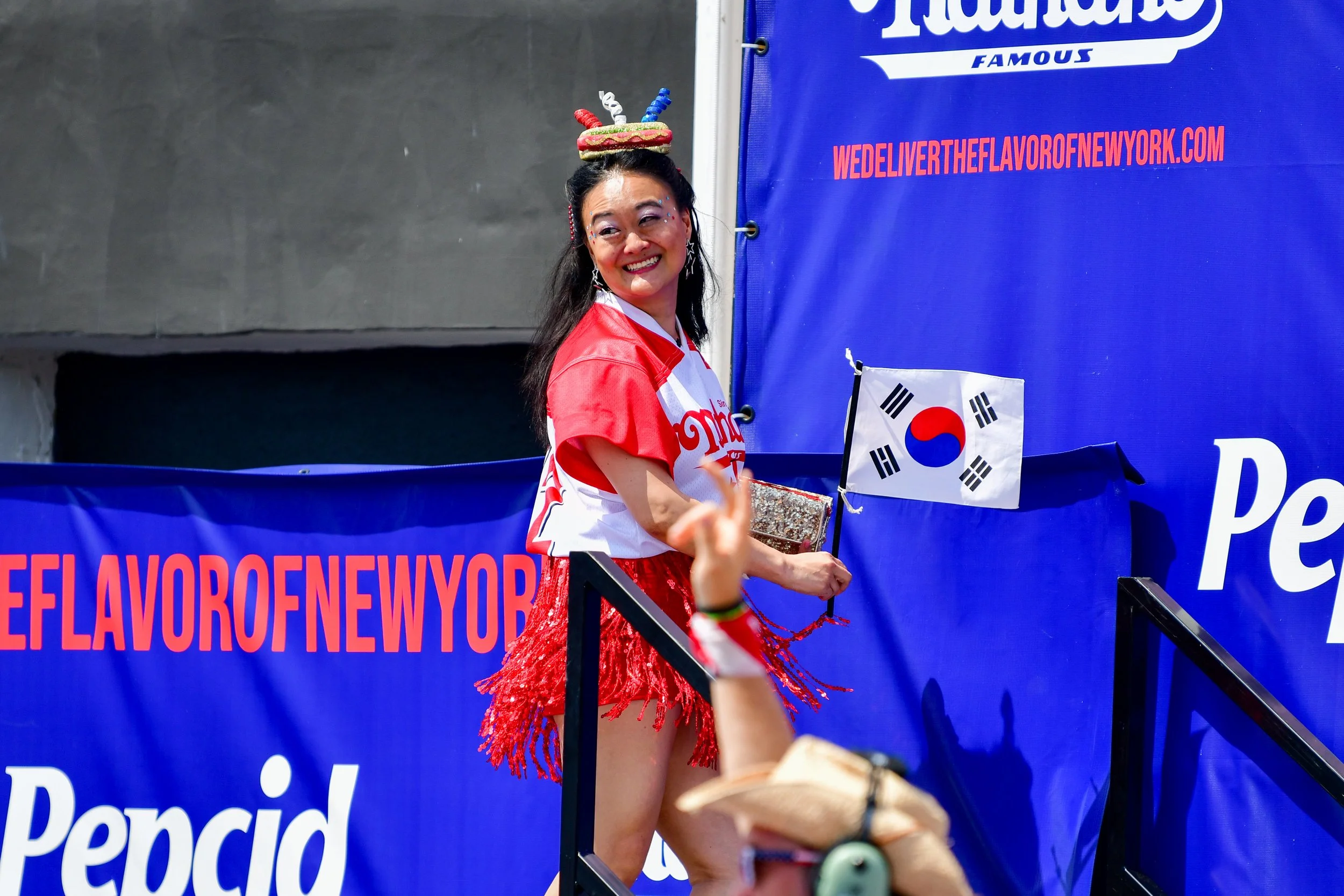 Mary Bowers smiles as she wears her signature headpiece resembling a hot dog at the 2023 Nathan's Famous International Hotdog Eating Contest. She is dressed in red and holding a glittery clutch and a Korean flag.