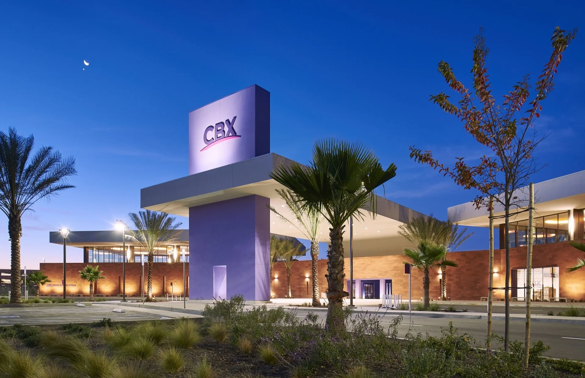 Building exterior at the Cross Border Xpress, with illuminated CBX sign on a tall purple structure, palm trees, and landscaped parking lot at twilight with a crescent moon and planets in the sky.