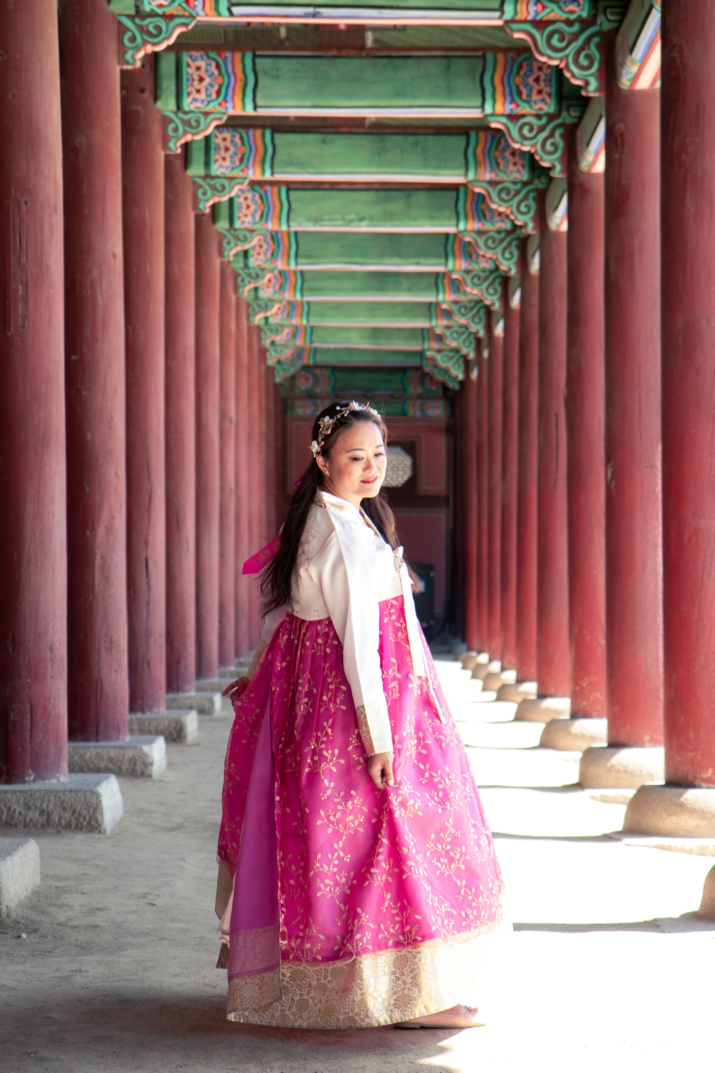 Mary Bowers dressed in traditional Korean hanbok, standing in a corridor with red wooden pillars and colorful, ornate ceiling details at Gyeongbokgung Palace.