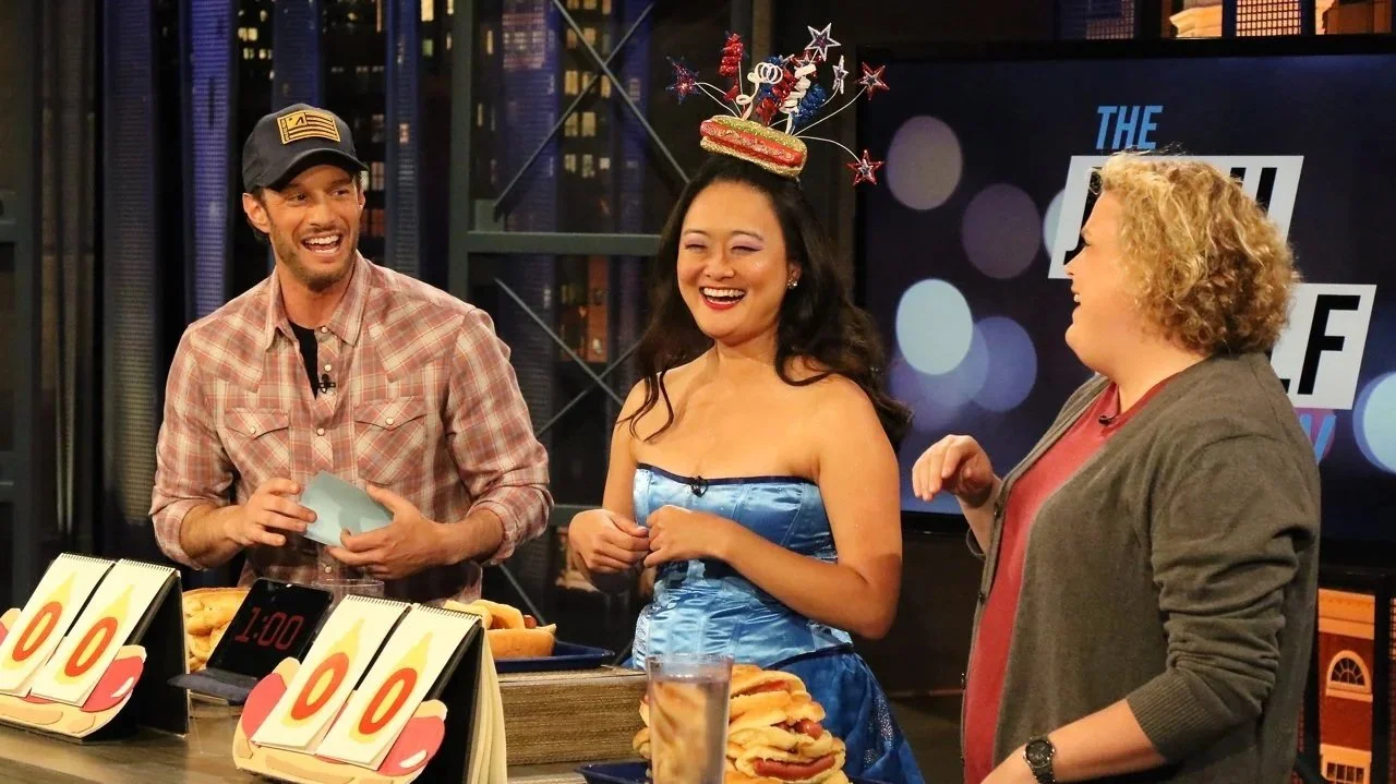 Mary Bowers wears her signature hotdog fascinator, as she laughs with Josh Wolf and Fortune Feimster on The Josh Wolf Show. The host and guests are surrounded by hotdogs and scorecards, as they prepare for an eating contest.