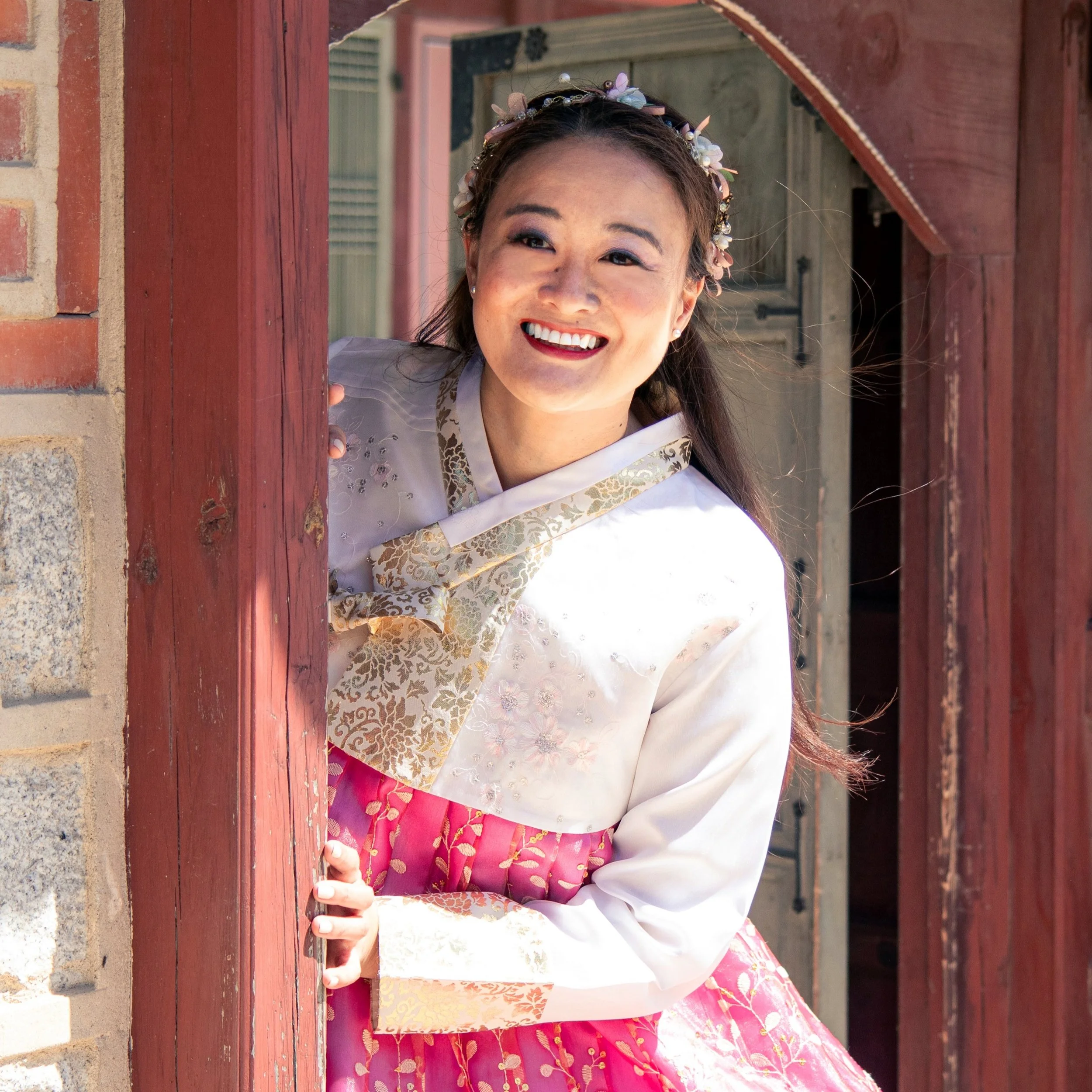 Mary Bowers wears a hanbok, as she leans out from a wooden window frame at Gyeongbokgung Palace, smiling.